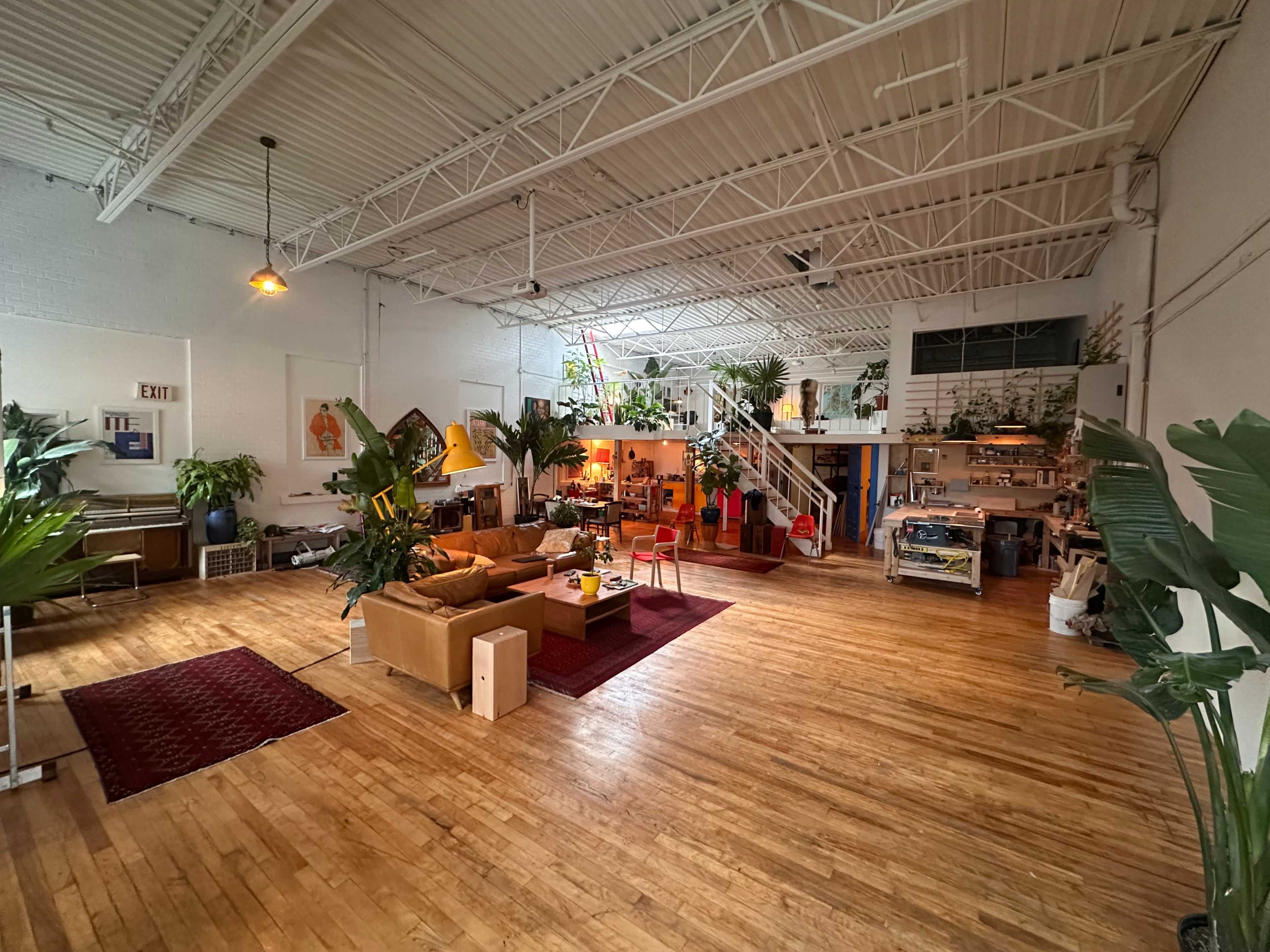 The image shows a spacious, open-plan studio or loft with wooden flooring, multiple potted plants, and various furniture arrangements, including a sofa and an area rug.