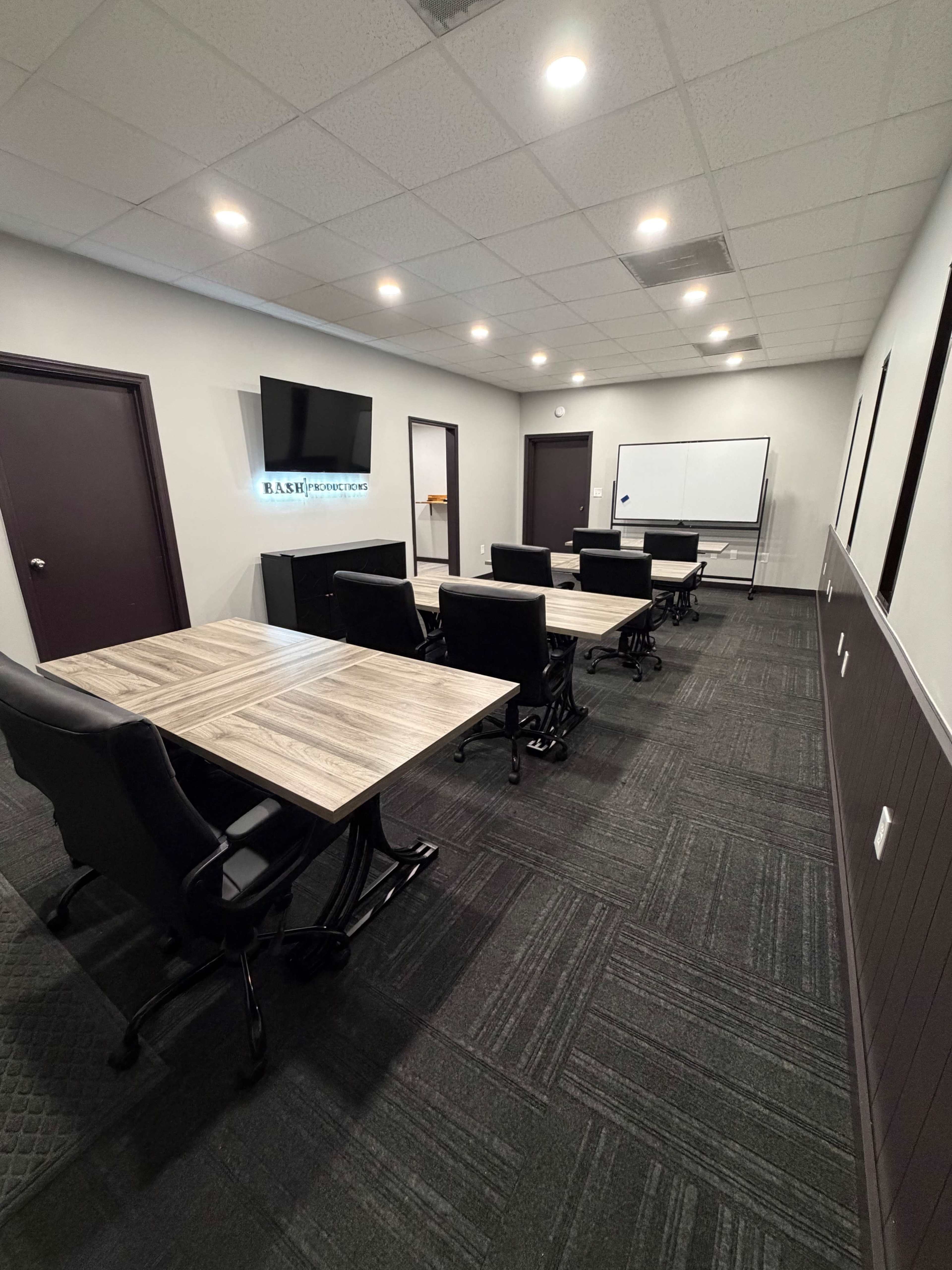 The image shows a modern conference room featuring several black office chairs around wooden tables, with a television and whiteboard mounted on the wall.