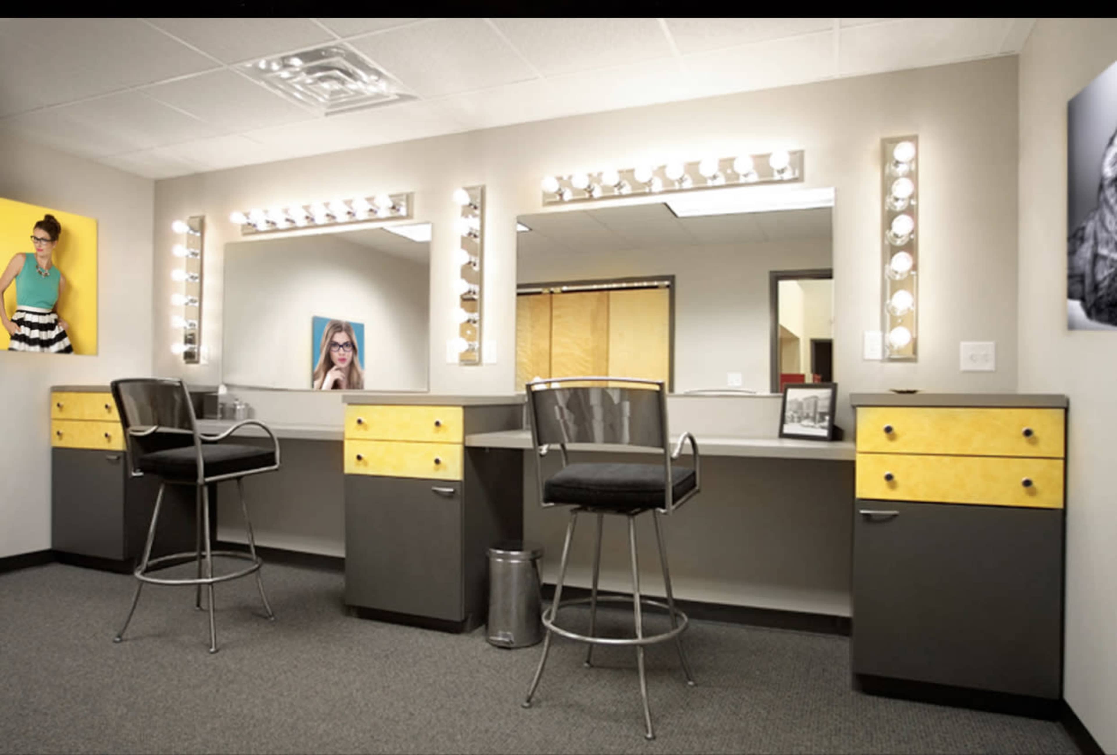 A well-lit dressing room featuring two vanity mirrors with multiple light bulbs, accompanied by high chairs and storage cabinets.