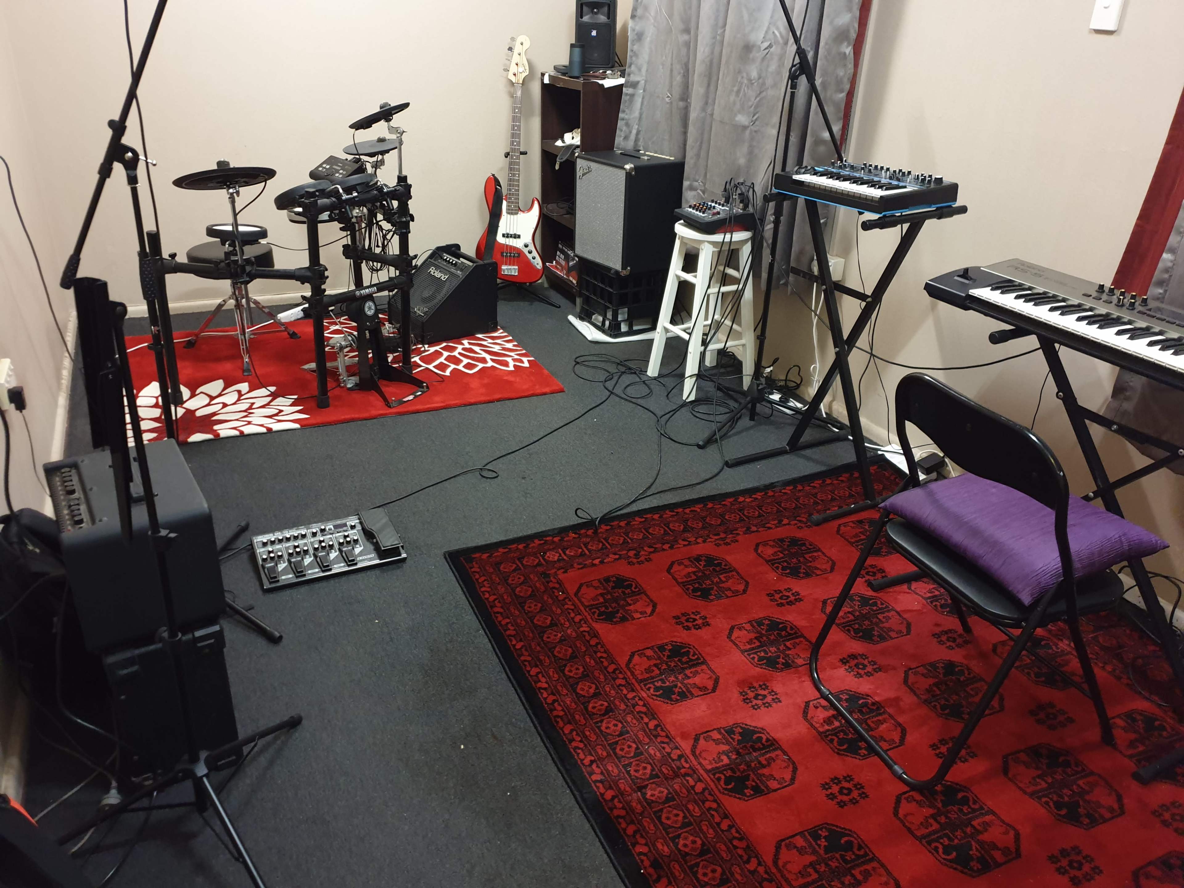 The room features various musical instruments, including an electric guitar, electronic drum set, synthesizer, and a keyboard, arranged on patterned rugs.