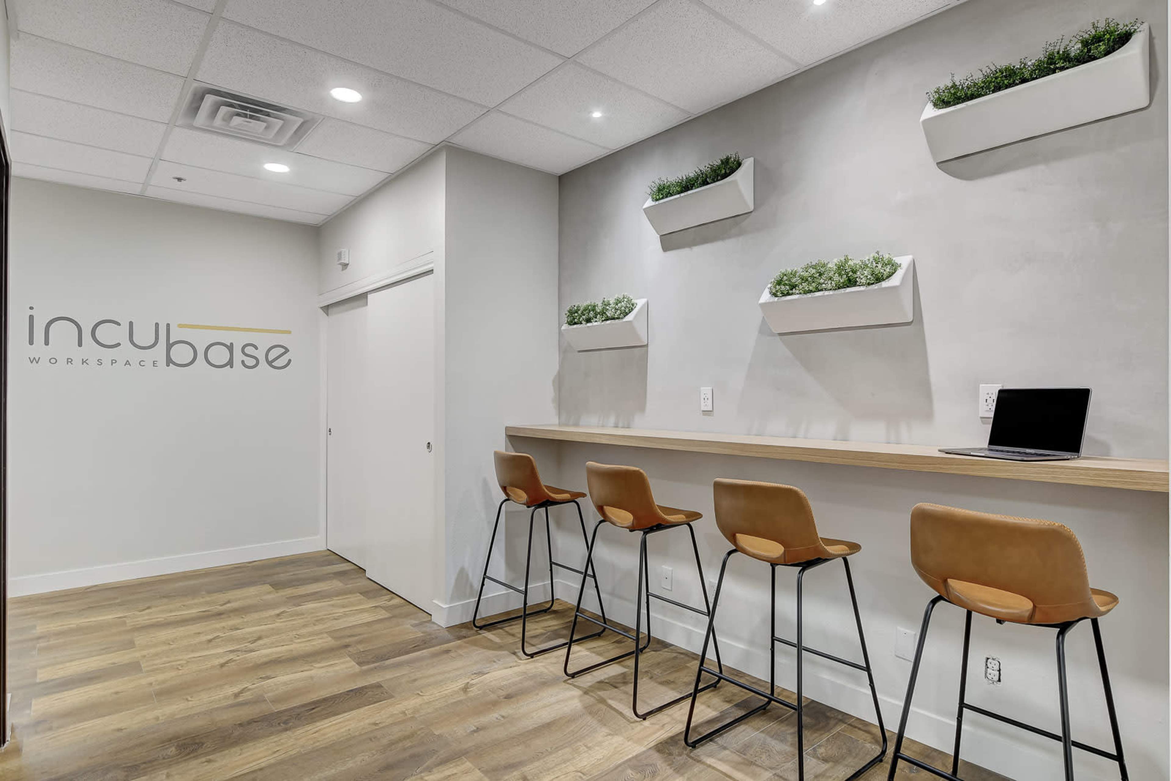 A modern workspace features a wooden counter with barstools, positioned against a wall adorned with planters and a logo for "incubase."