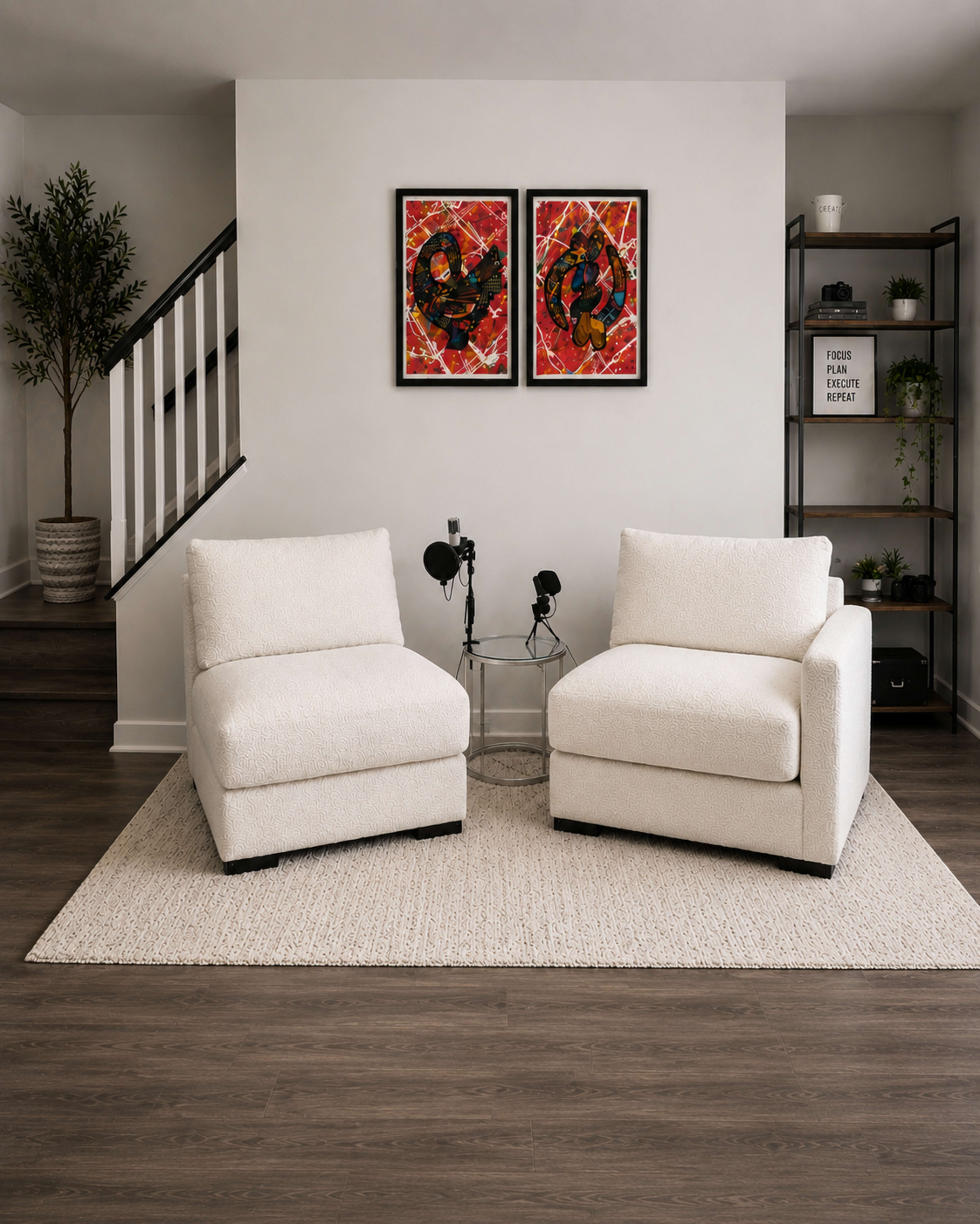 The image shows a modern living room featuring two white chairs, a small table between them, and colorful artwork on the wall, with a plant and shelving unit in the background.