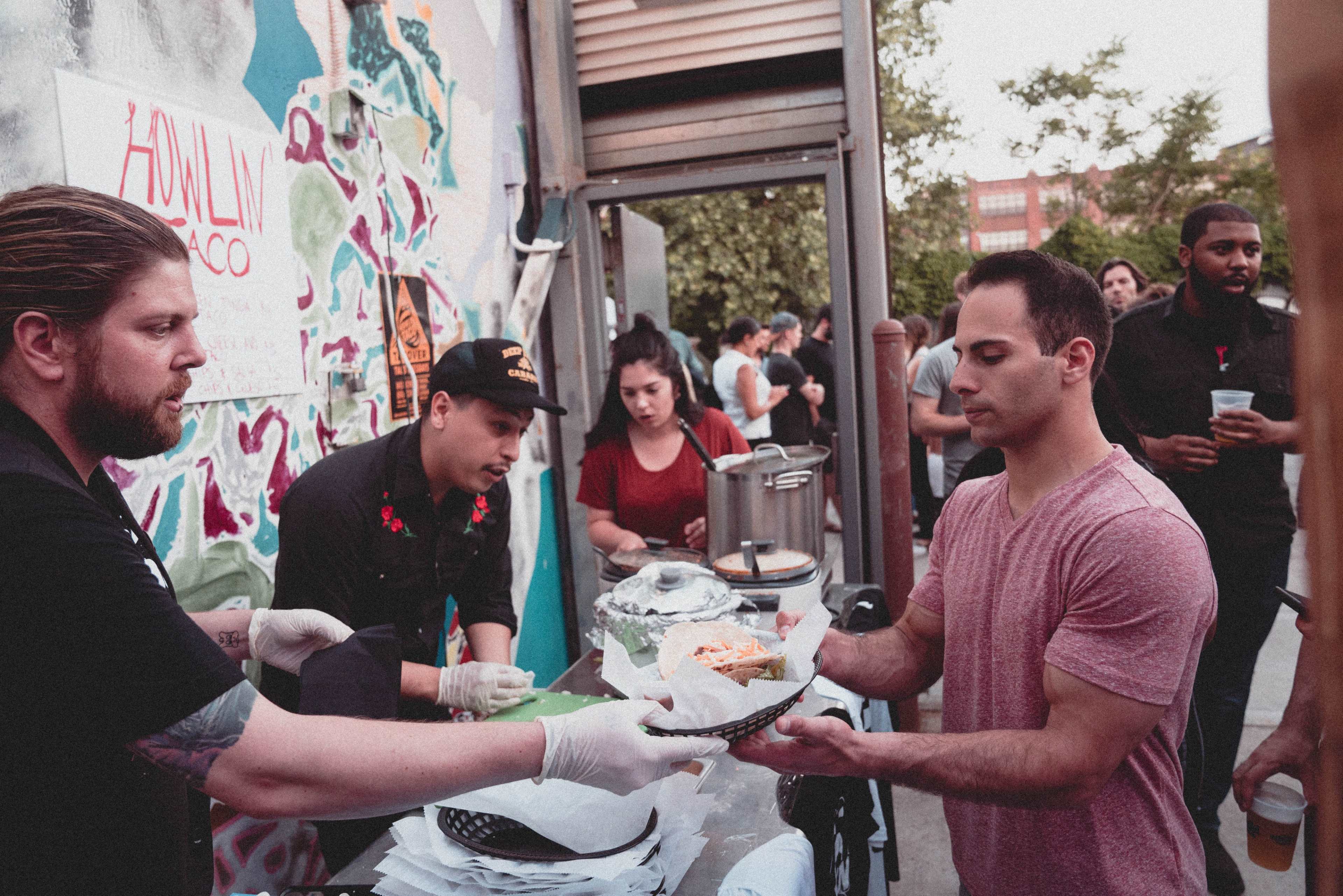 A group of people at an outdoor event are serving food from a counter to attendees, while others wait in line.
