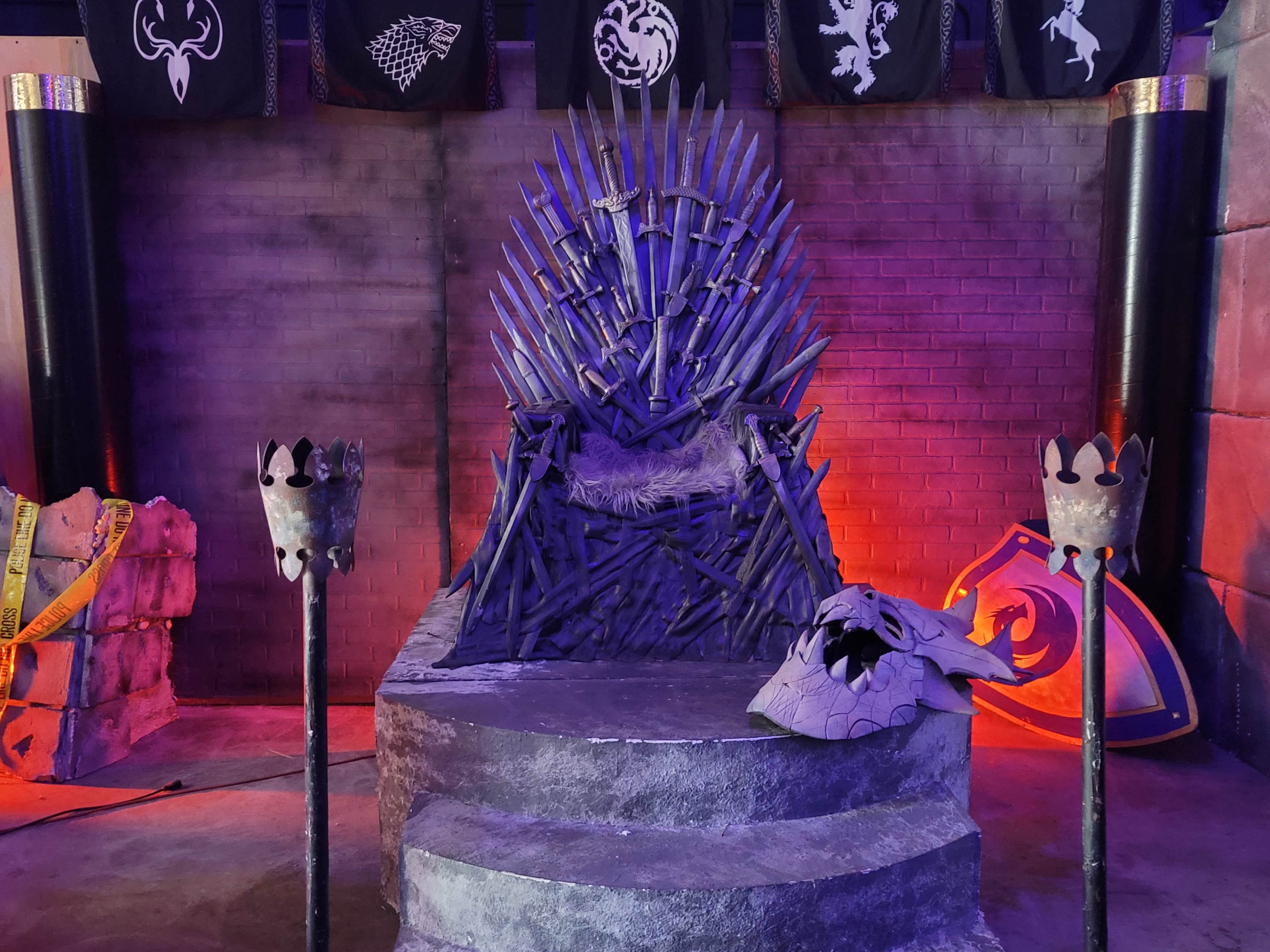 The image shows a replica of a throne made of swords, set against a backdrop of banners and illuminated by colored lights.