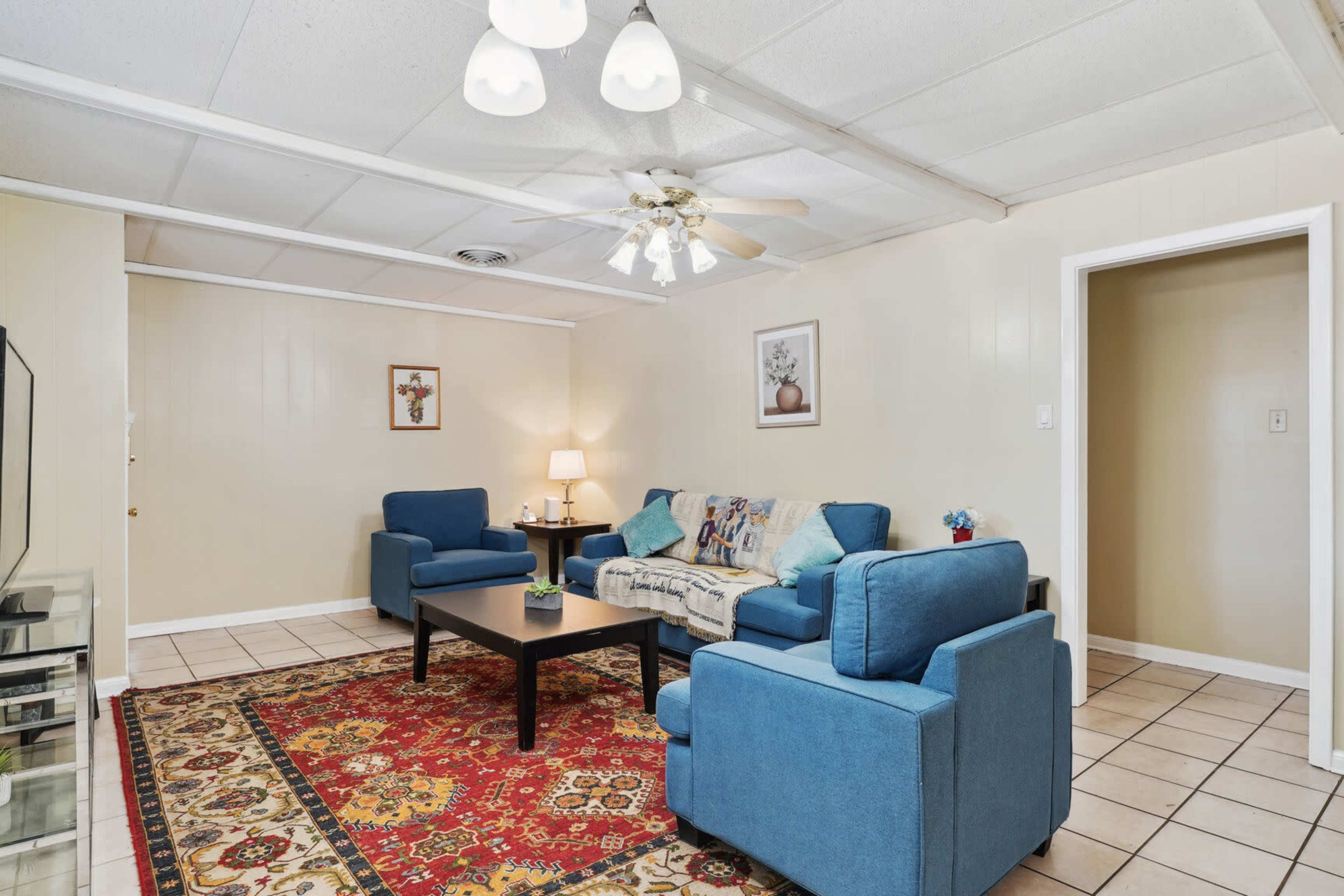 The image shows a living room with two blue couches, a coffee table, a decorative rug, and a ceiling fan, along with a lamp and wall art.