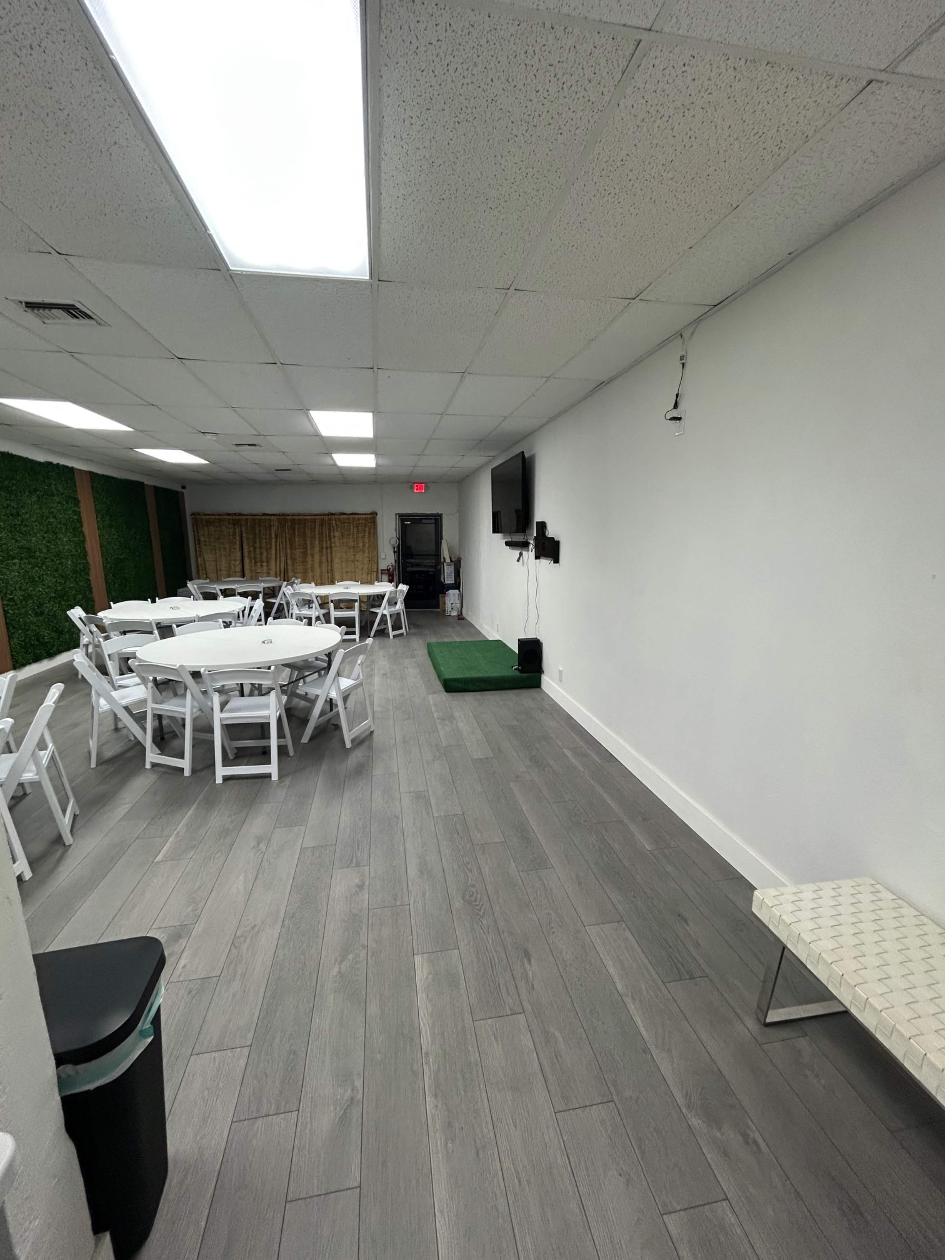 The image shows a spacious indoor venue with white folding tables and chairs arranged in a seating configuration, a television mounted on the wall, and a small stage area with a green carpet.