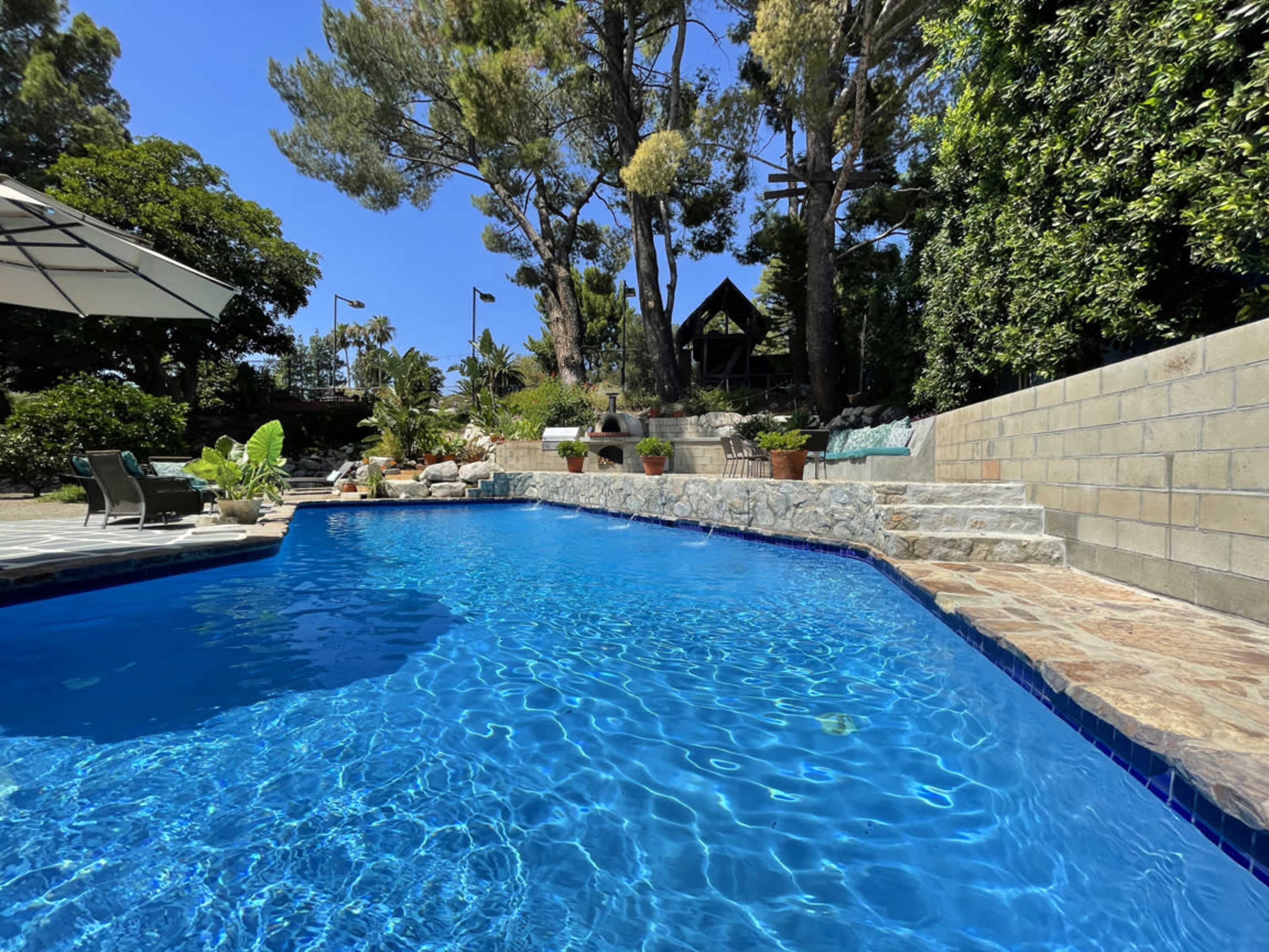 A clear blue swimming pool surrounded by stone steps, greenery, and tall trees under a bright sky.