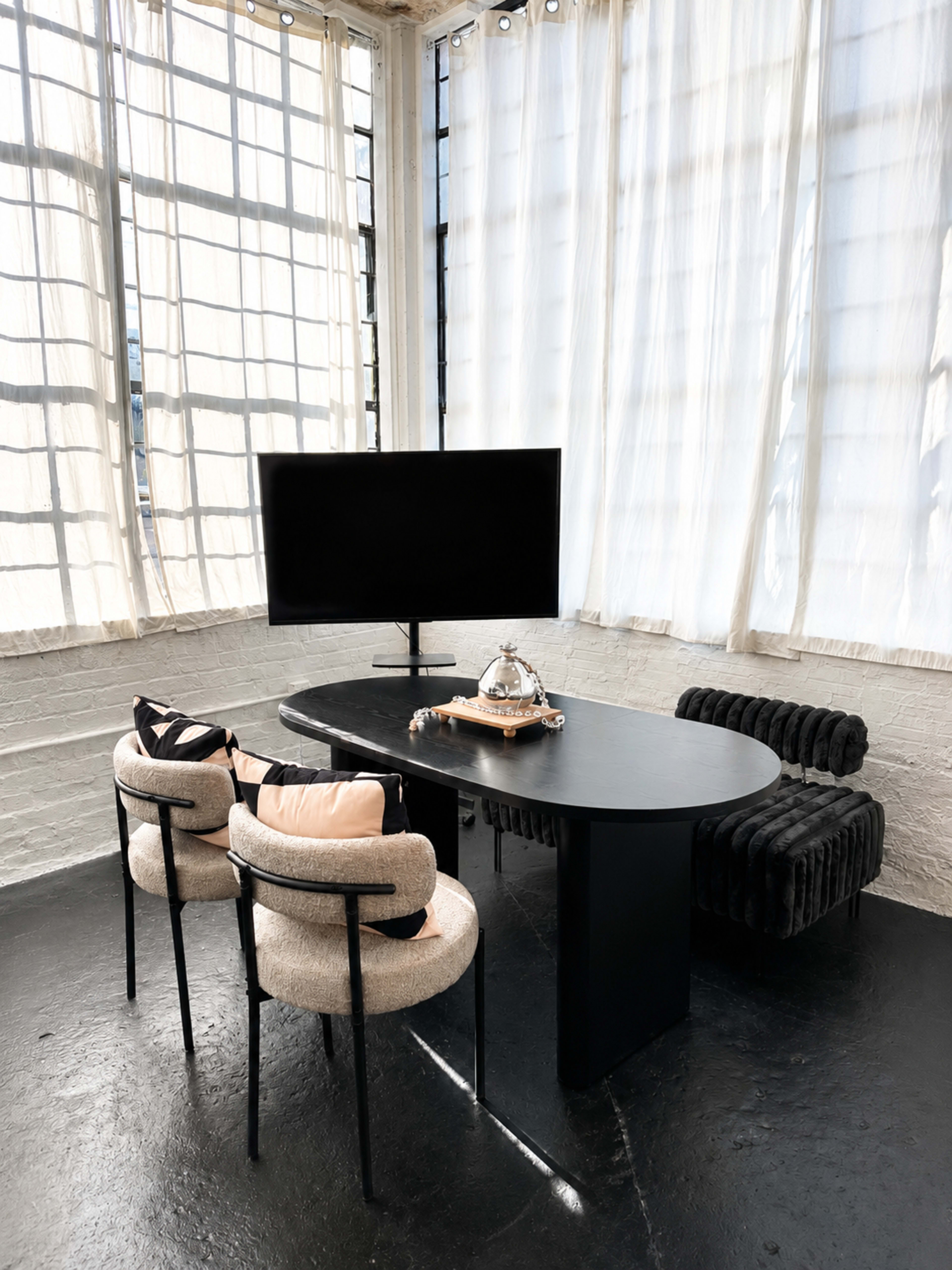 A minimalist workspace features a large black table with two chairs and a television, positioned next to large windows with sheer curtains.