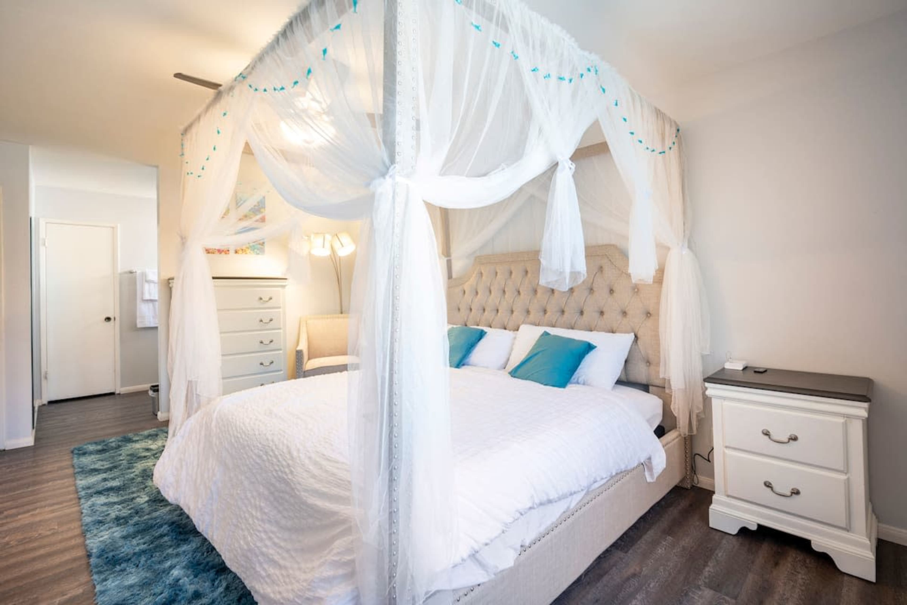 A canopy bed with sheer drapes and decorative accents is positioned in a well-lit bedroom featuring a nightstand and a chair nearby.