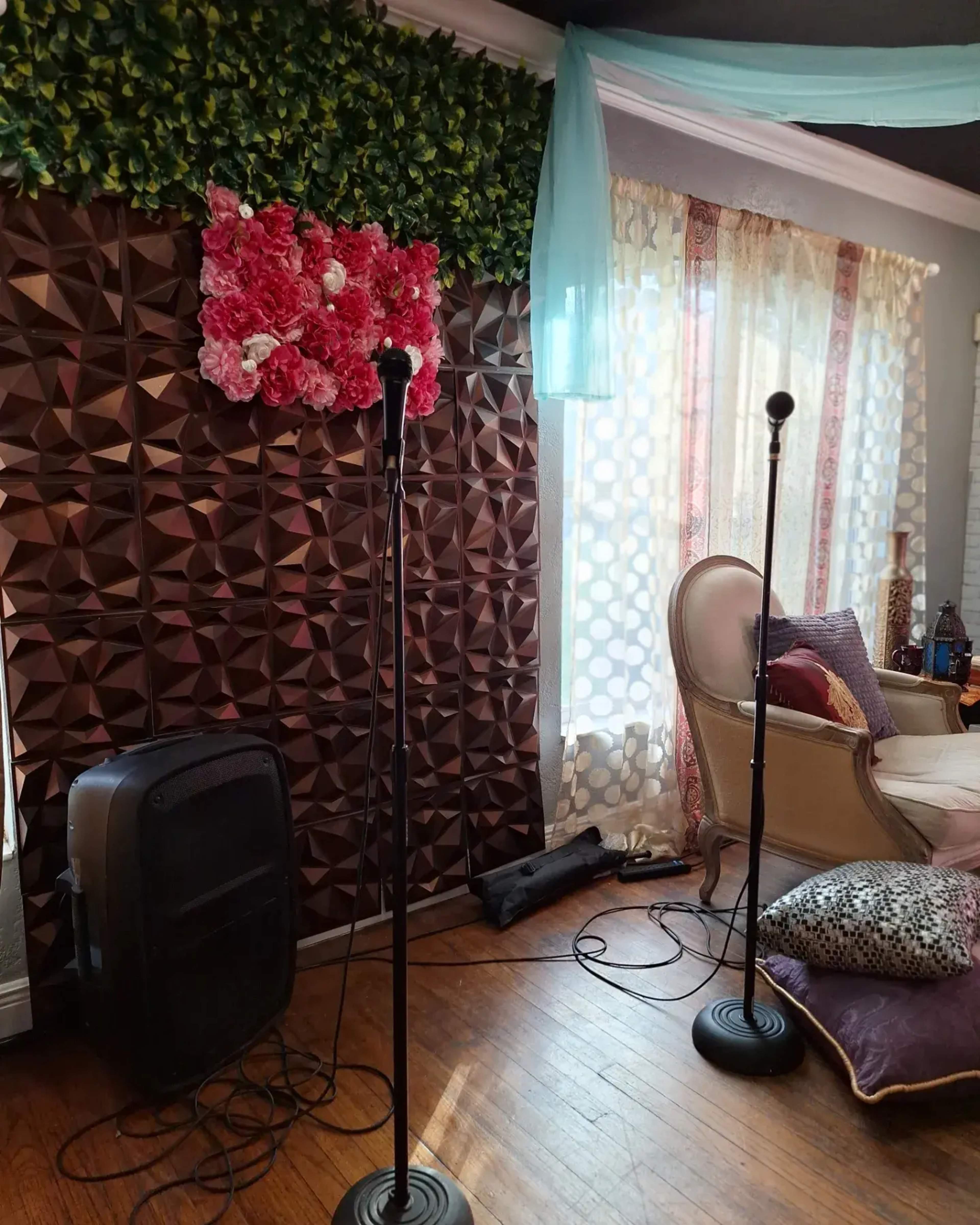A room with two microphone stands positioned in front of a decorative wall covered in brown geometric panels and a floral arrangement, with large windows draped in sheer curtains.