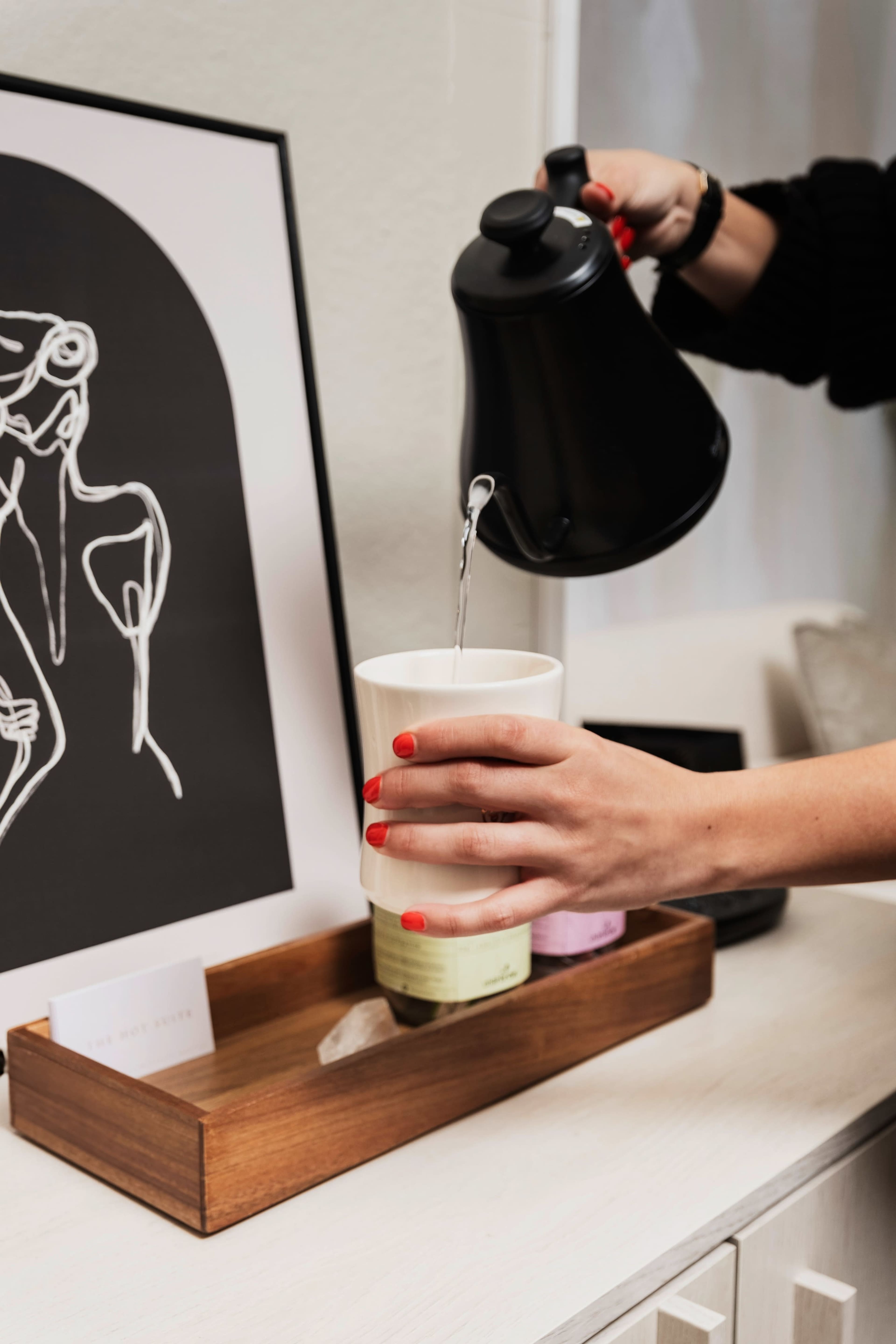A person pours water from a black kettle into a white cup placed on a wooden tray next to an art print featuring a line drawing.