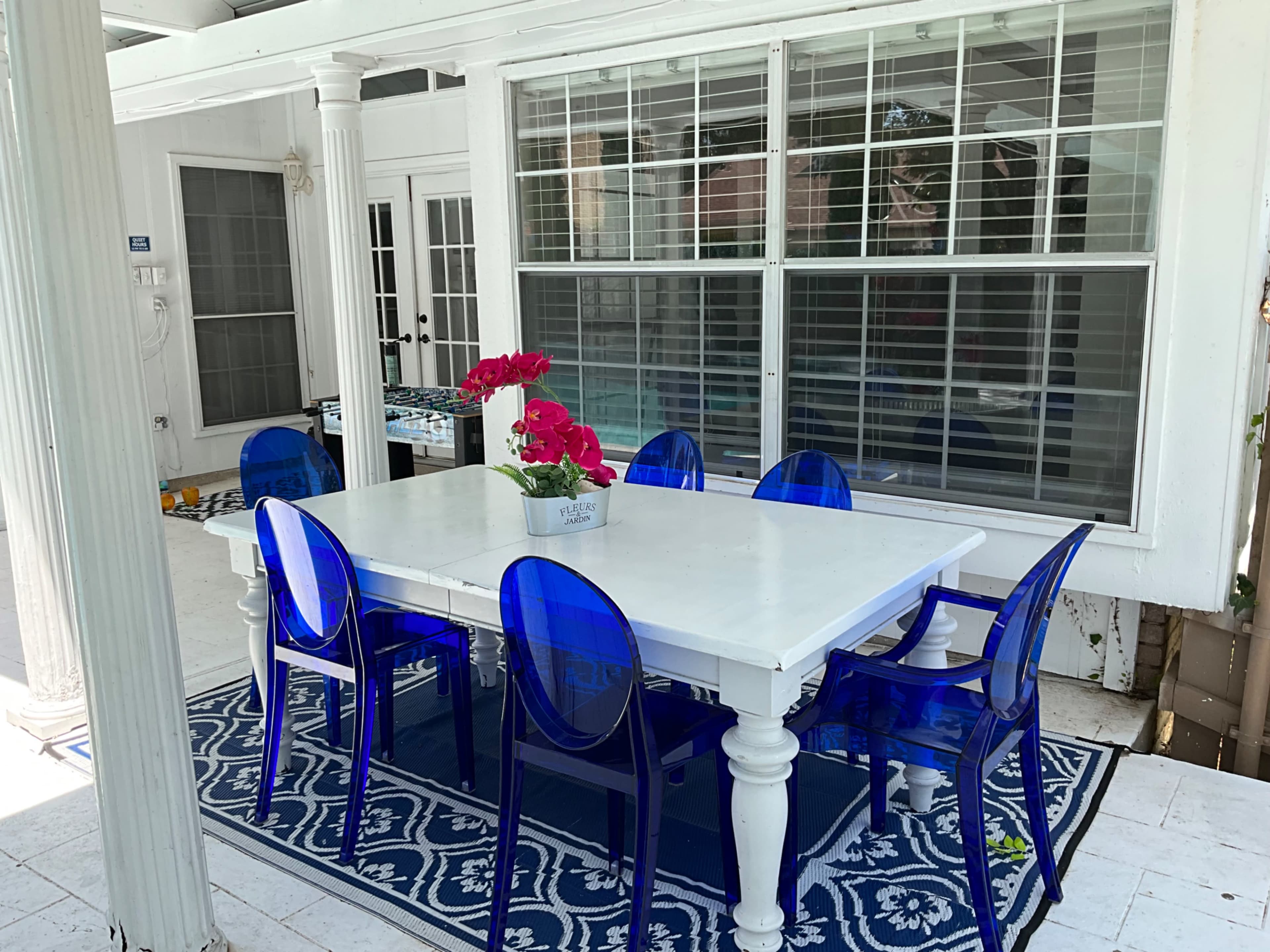 A white dining table surrounded by blue transparent chairs is set on a patterned rug in a sunlit patio area.