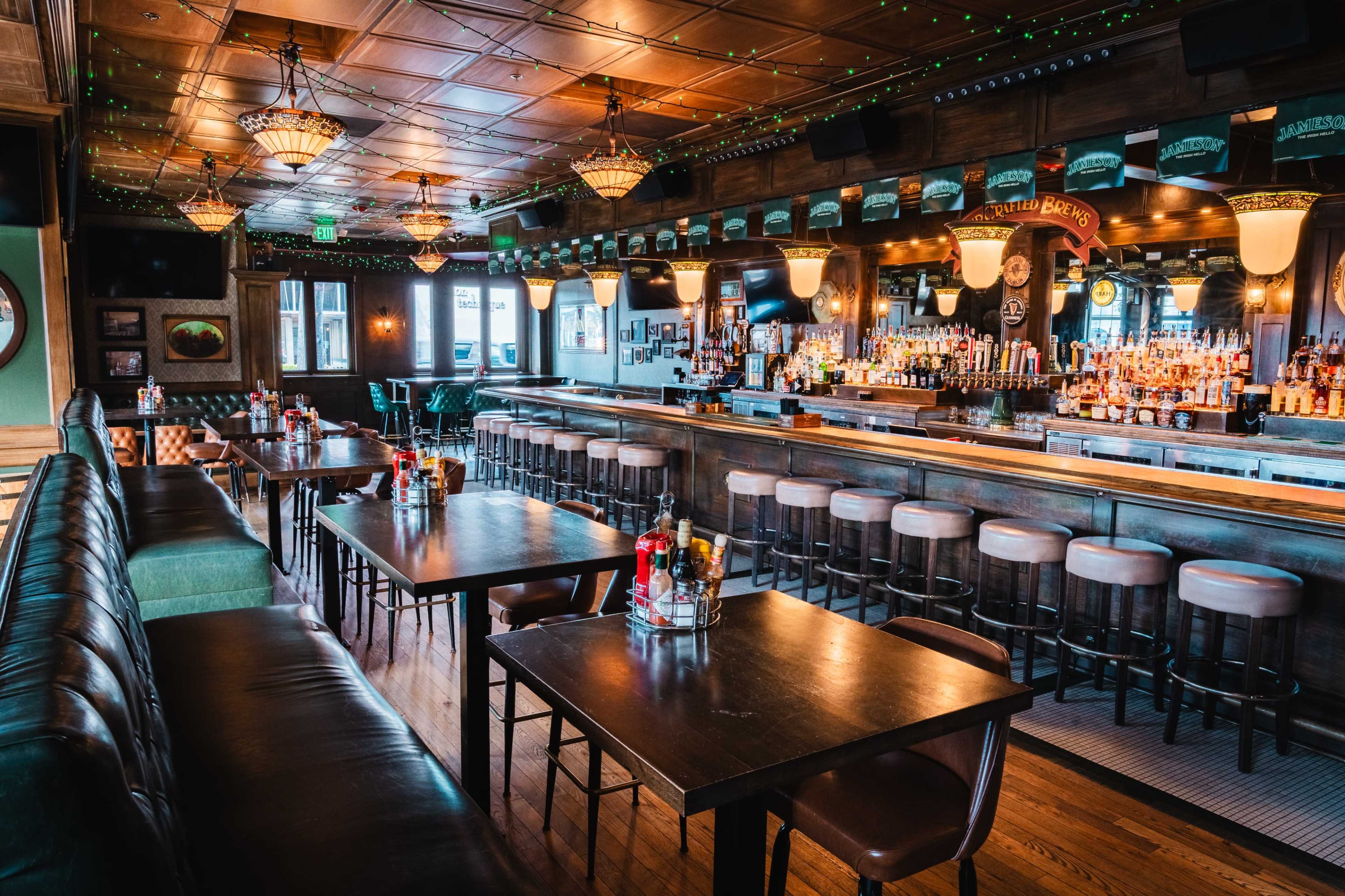 The image shows the interior of a bar with wooden finishes, a long counter lined with stools, and tables arranged for seating.