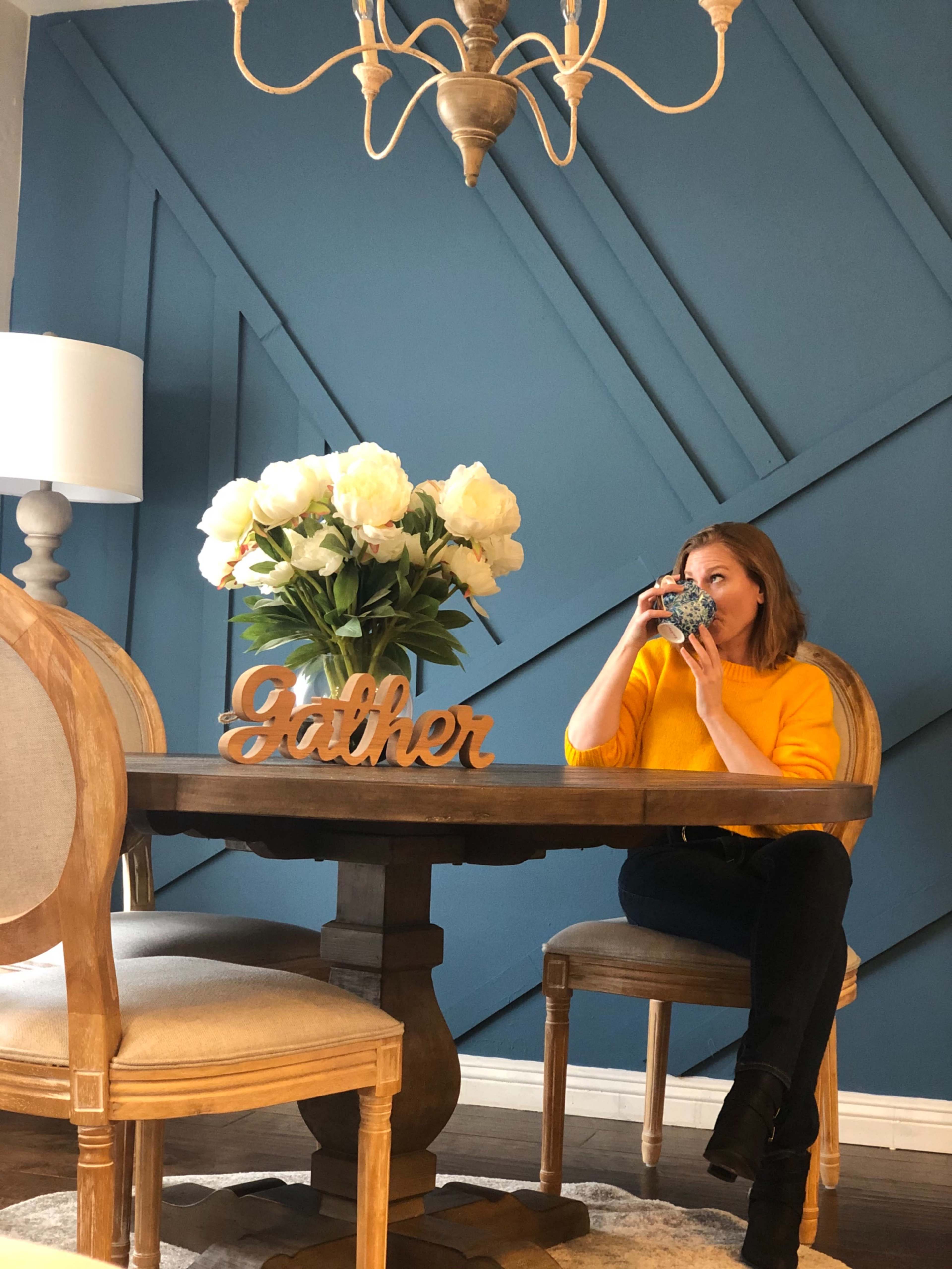 A woman in a yellow sweater sips from a mug at a dining table adorned with a floral centerpiece and a decorative "Gather" sign against a blue wall.