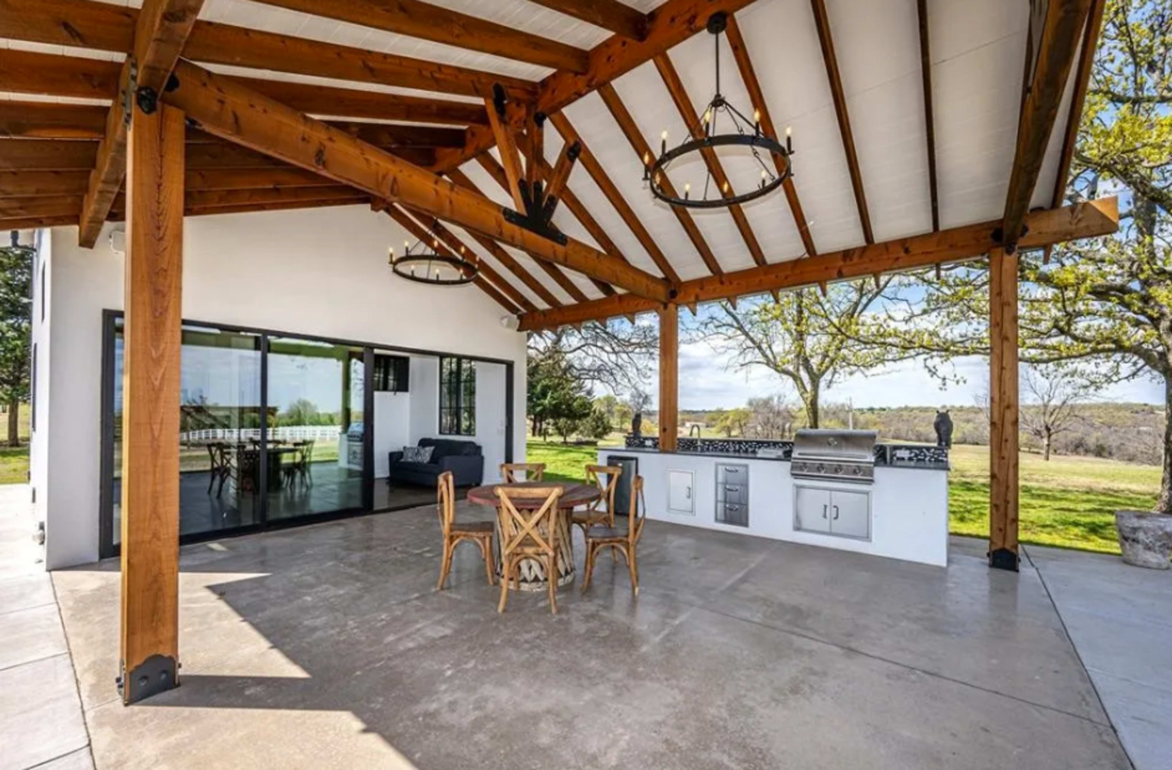 The image shows a covered outdoor patio with wooden beams, a dining table, and a built-in grill.