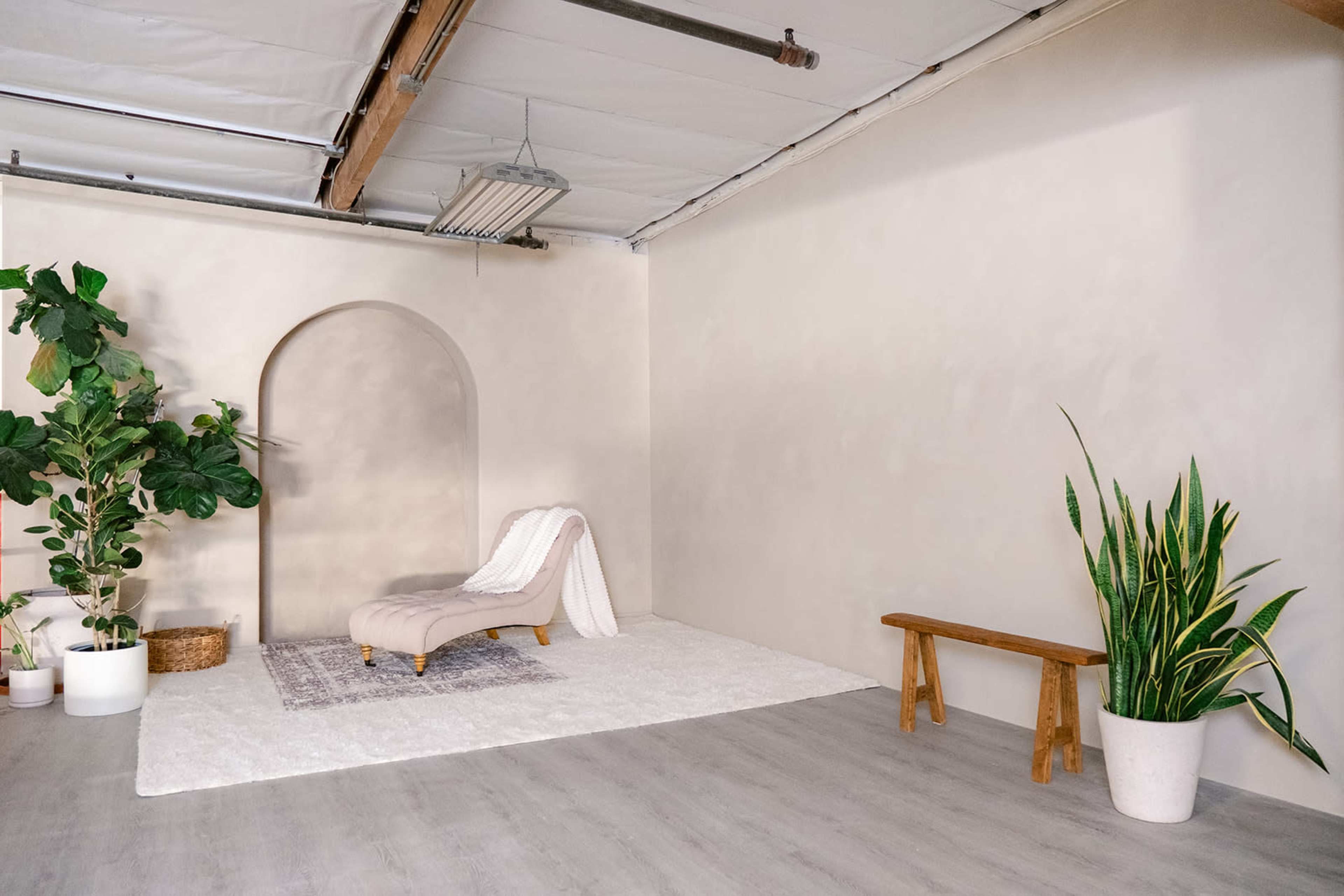A minimalist room features a light gray wall, a curved archway, a lounge chair, a wooden bench, and potted plants.