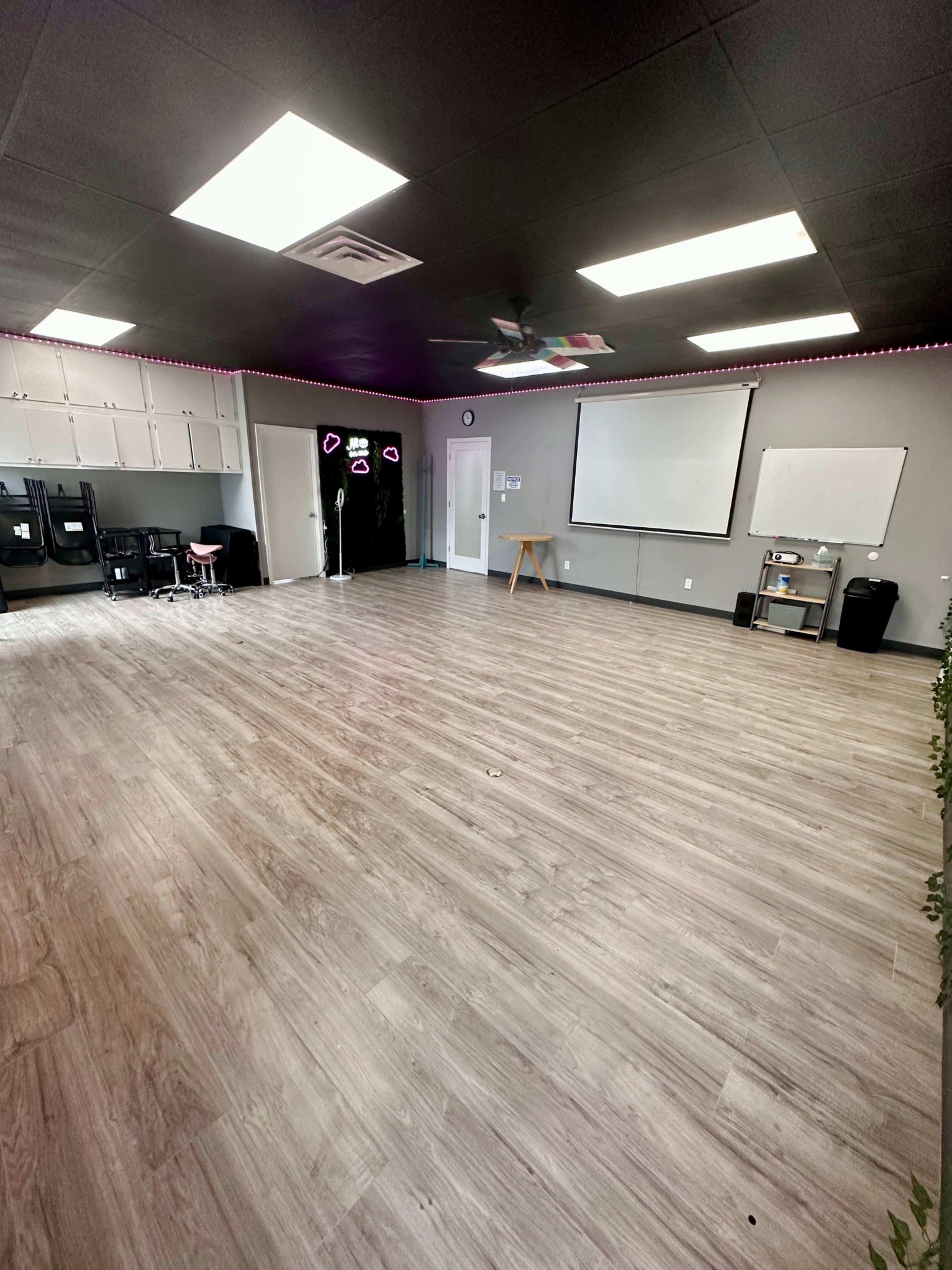 An empty room with wooden flooring, white walls, and various presentation screens at the front, along with chairs and a small table positioned in the corner.