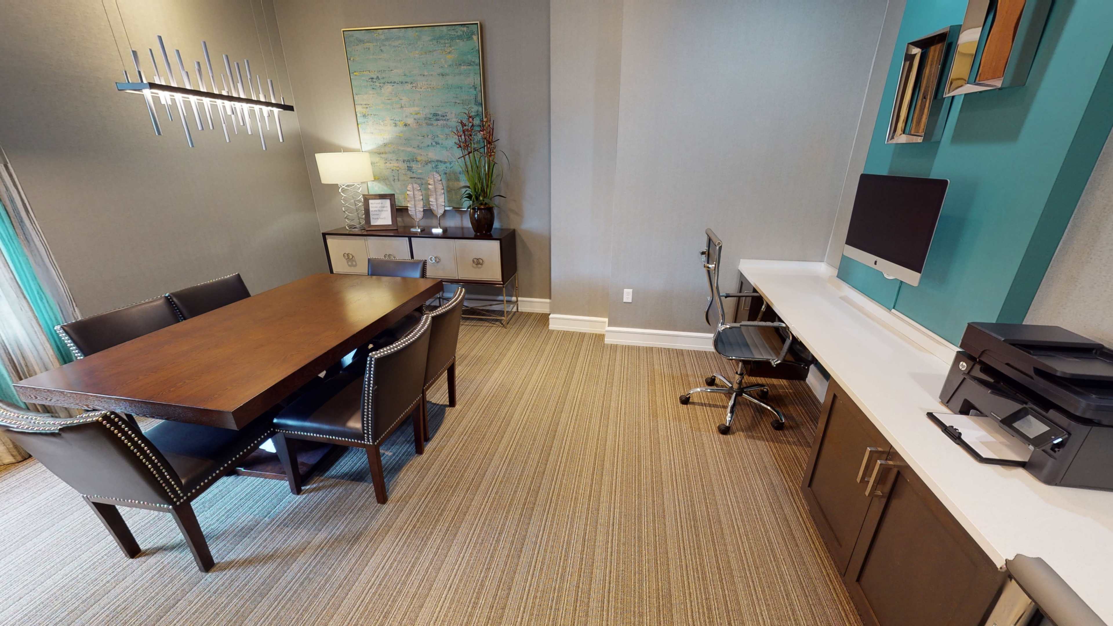 A modern office space featuring a large wooden dining table surrounded by chairs, a computer desk with an Apple monitor, and a printer nearby, all set against a neutral-toned wall.
