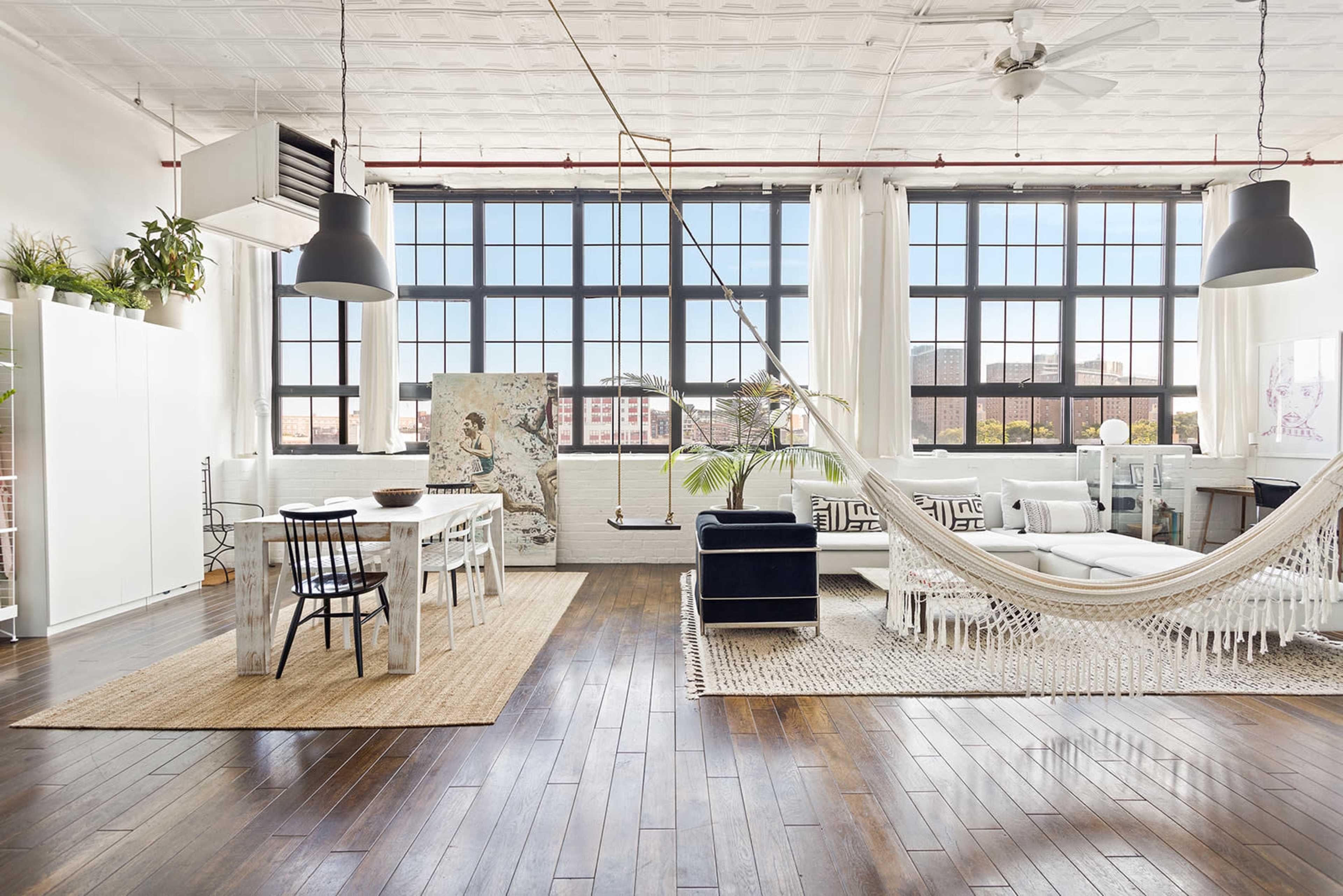 A spacious, well-lit room features a dining area with a table and chairs, a hammock, and a seating area with a sofa, large windows, and hardwood flooring.