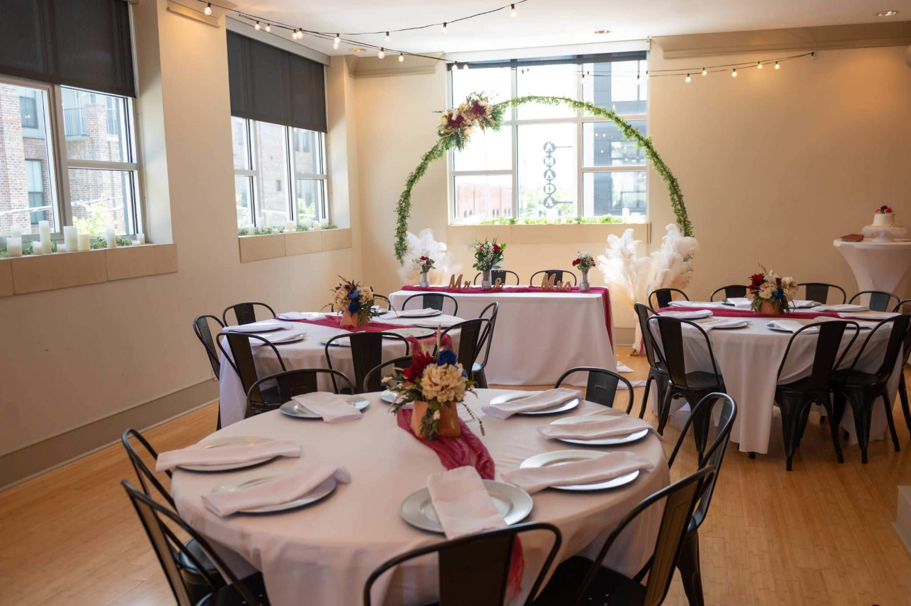 A spacious event room set up for a gathering, featuring round tables with white tablecloths, black chairs, and floral centerpieces, alongside a decorated serving table and large windows.