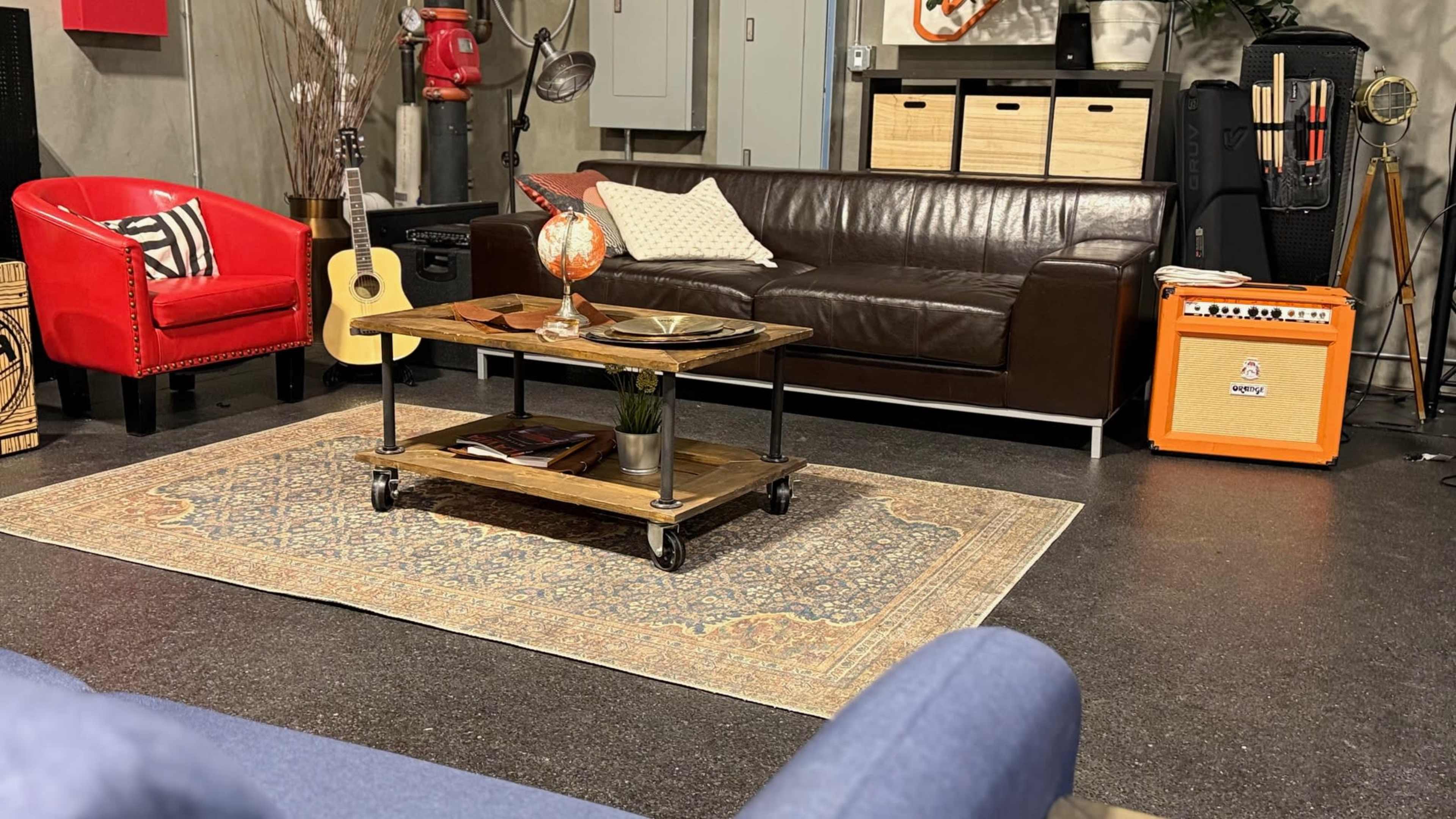 The scene features a cozy living area with a brown leather sofa, a wooden coffee table on wheels, a red armchair, an acoustic guitar, and an orange amplifier against a textured wall.