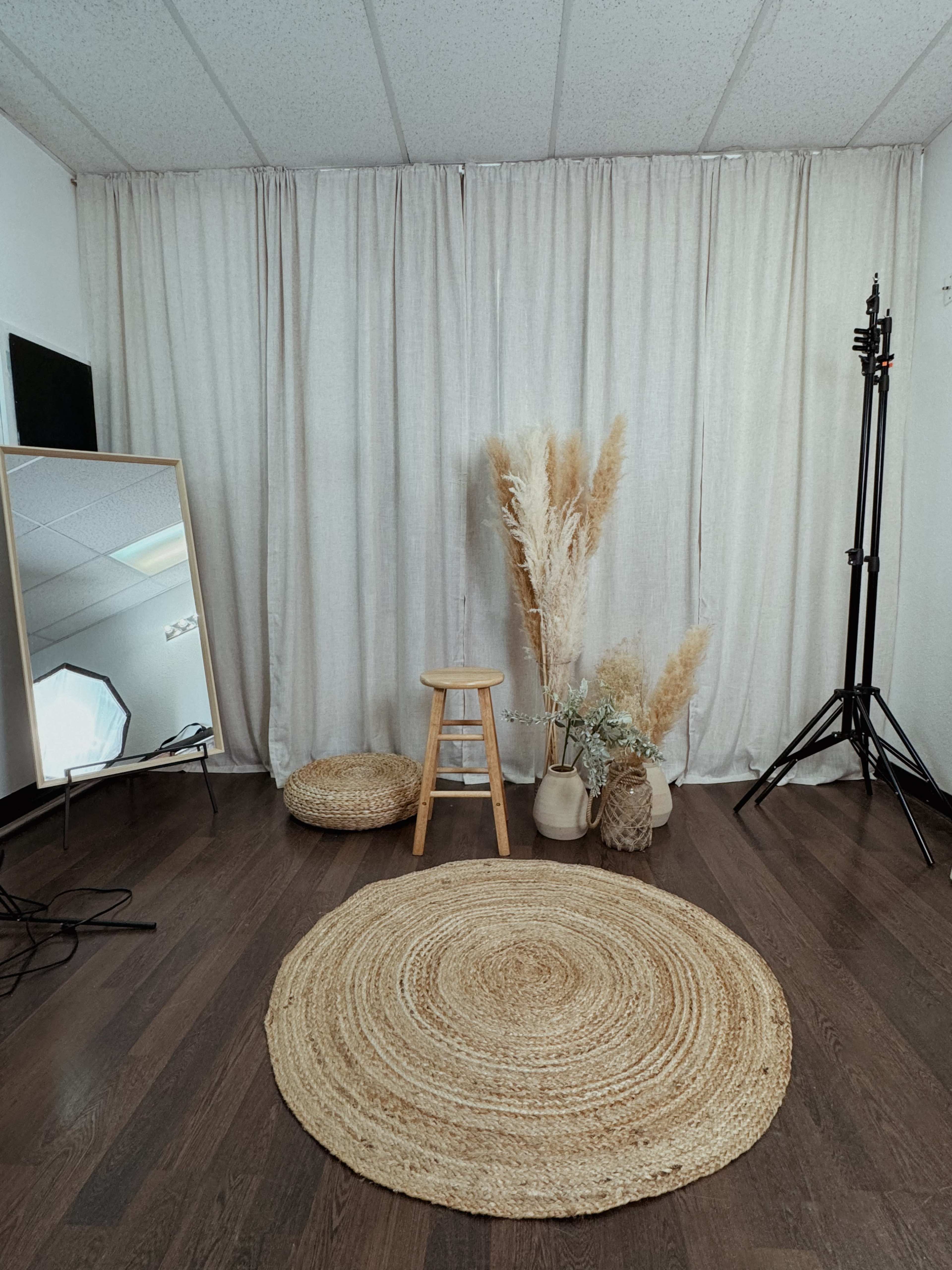 A minimalistic studio space featuring a wooden stool, a large mirror, a round jute rug, and decorative dried plants in vases, against a backdrop of light curtains.