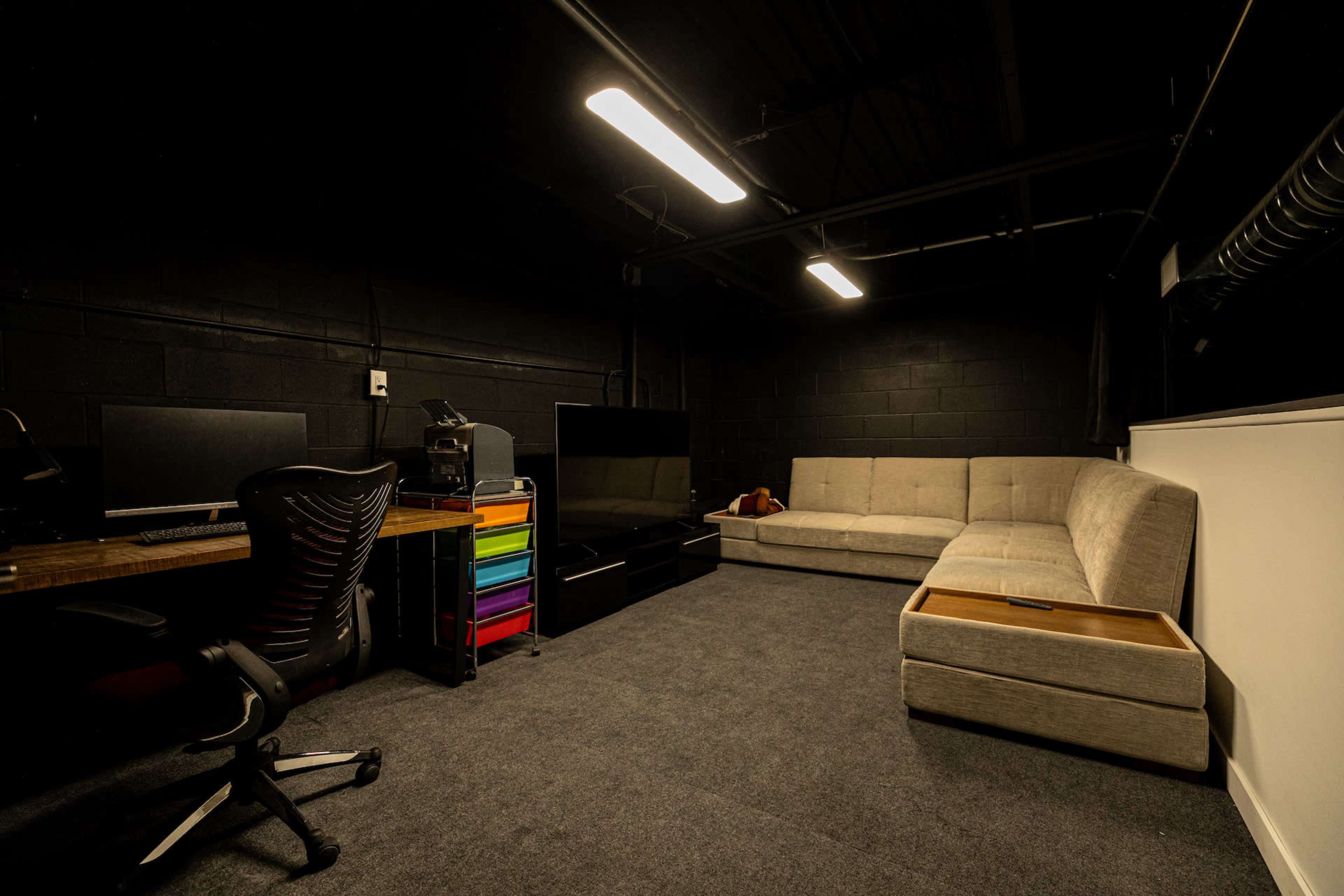 A dimly lit room features a desk with two computer monitors, a couch, and a multicolored storage unit against the wall.