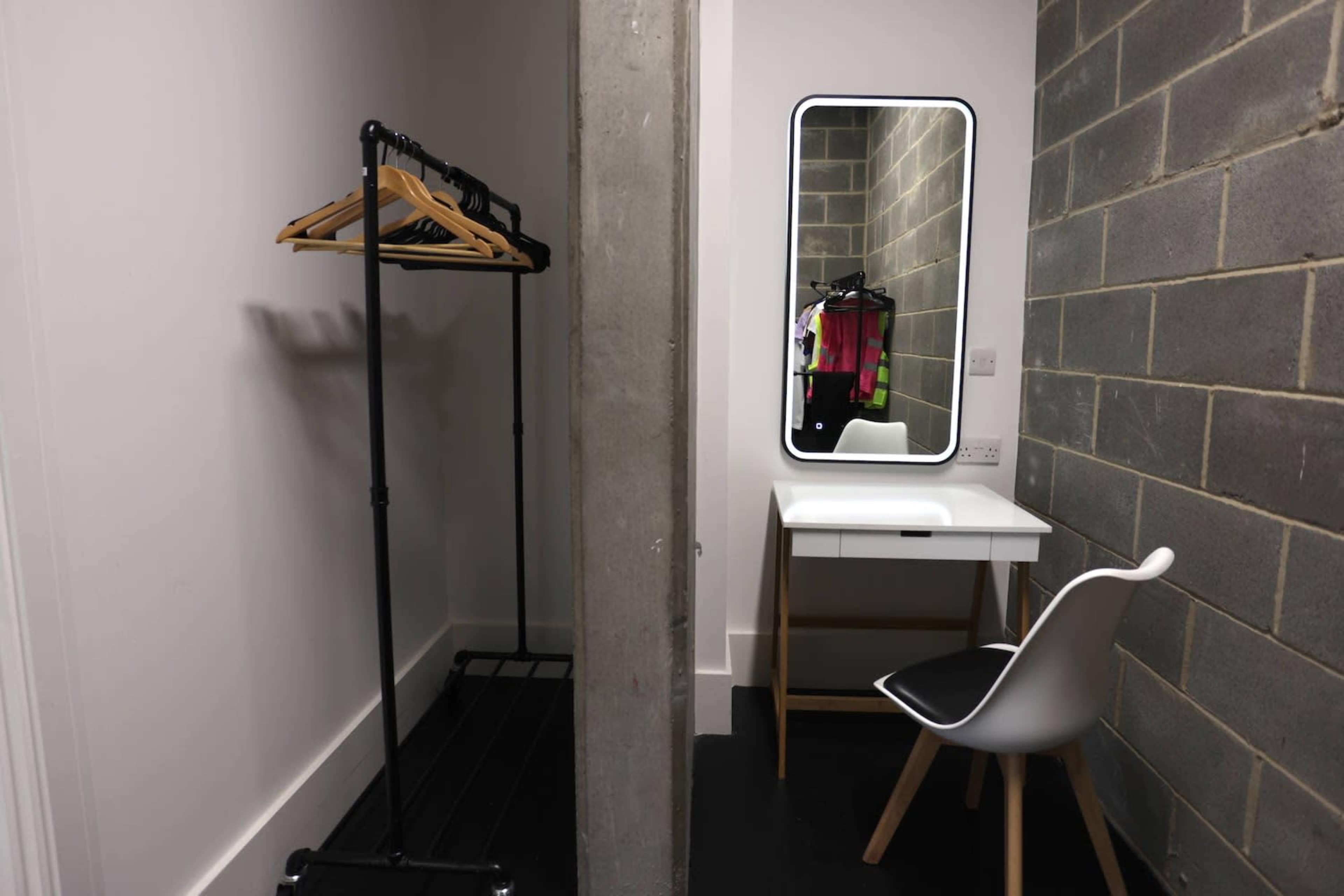A minimalistic dressing room with a clothing rack, a mirror, a desk, and a chair against a concrete wall.