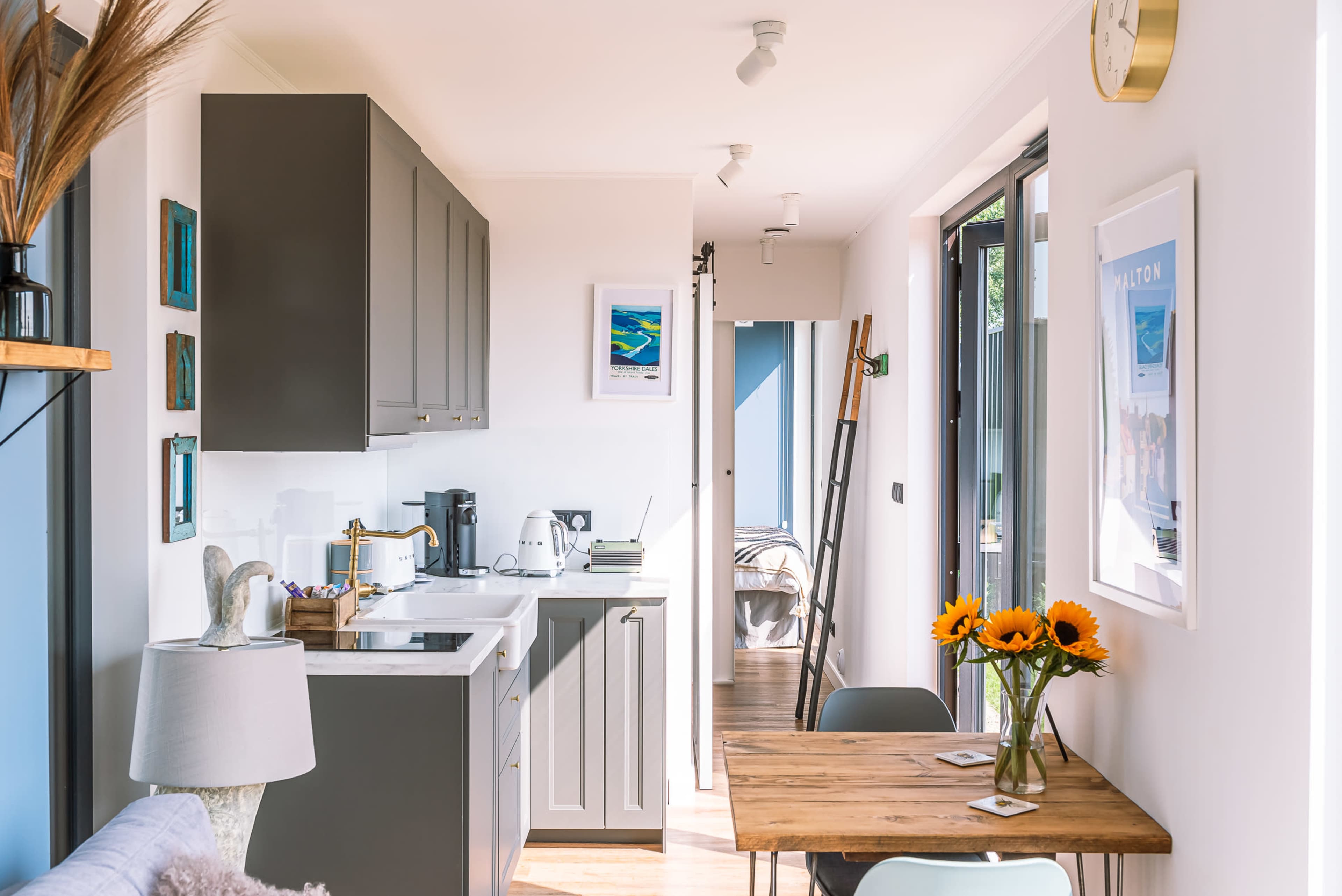 A modern kitchen and dining area feature gray cabinets, a small table with sunflowers, and natural light from large windows.