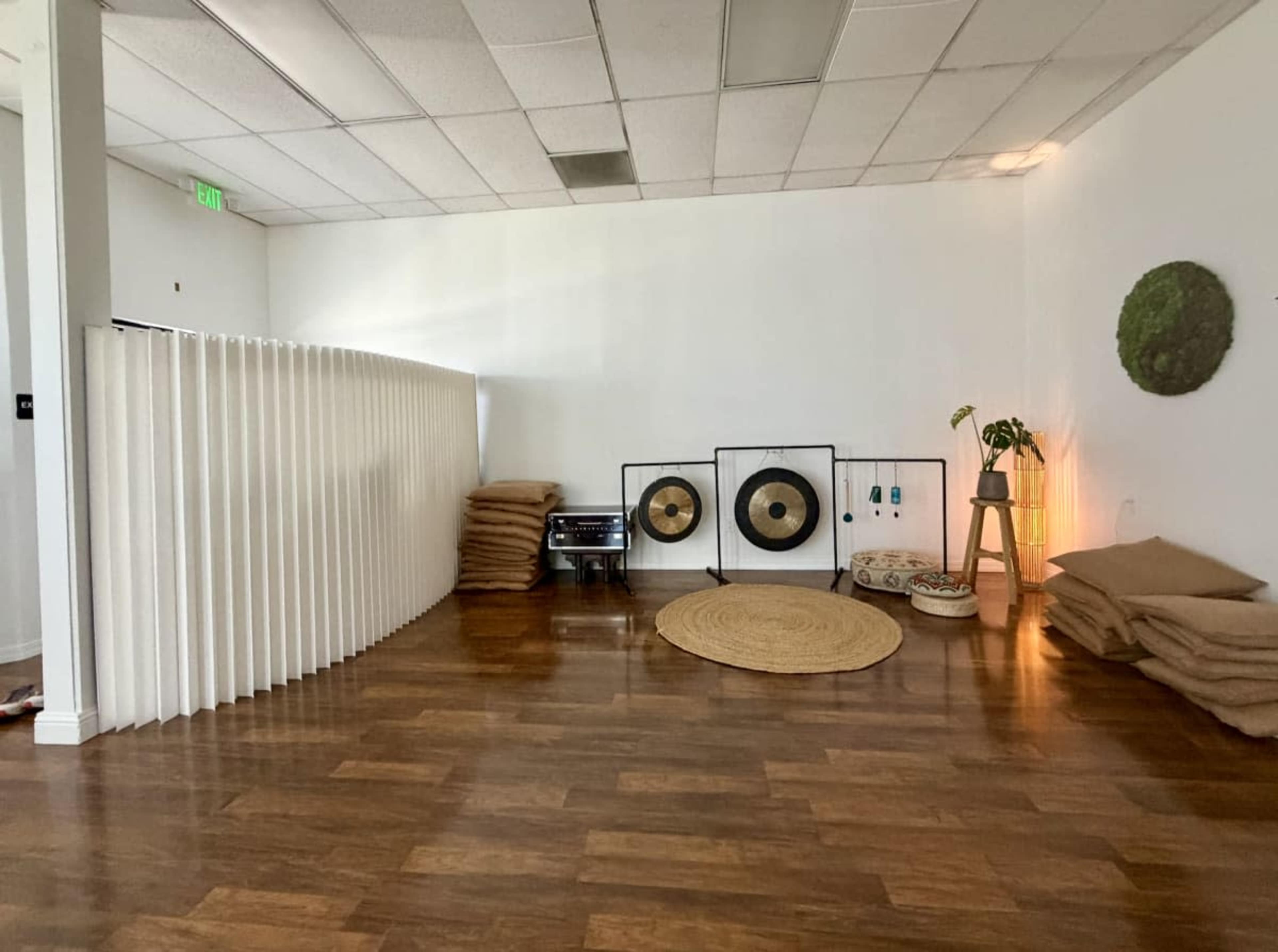 The image shows a spacious, well-lit room with wooden flooring, featuring a folding partition, circular rugs, large gongs, and stacks of cushions.
