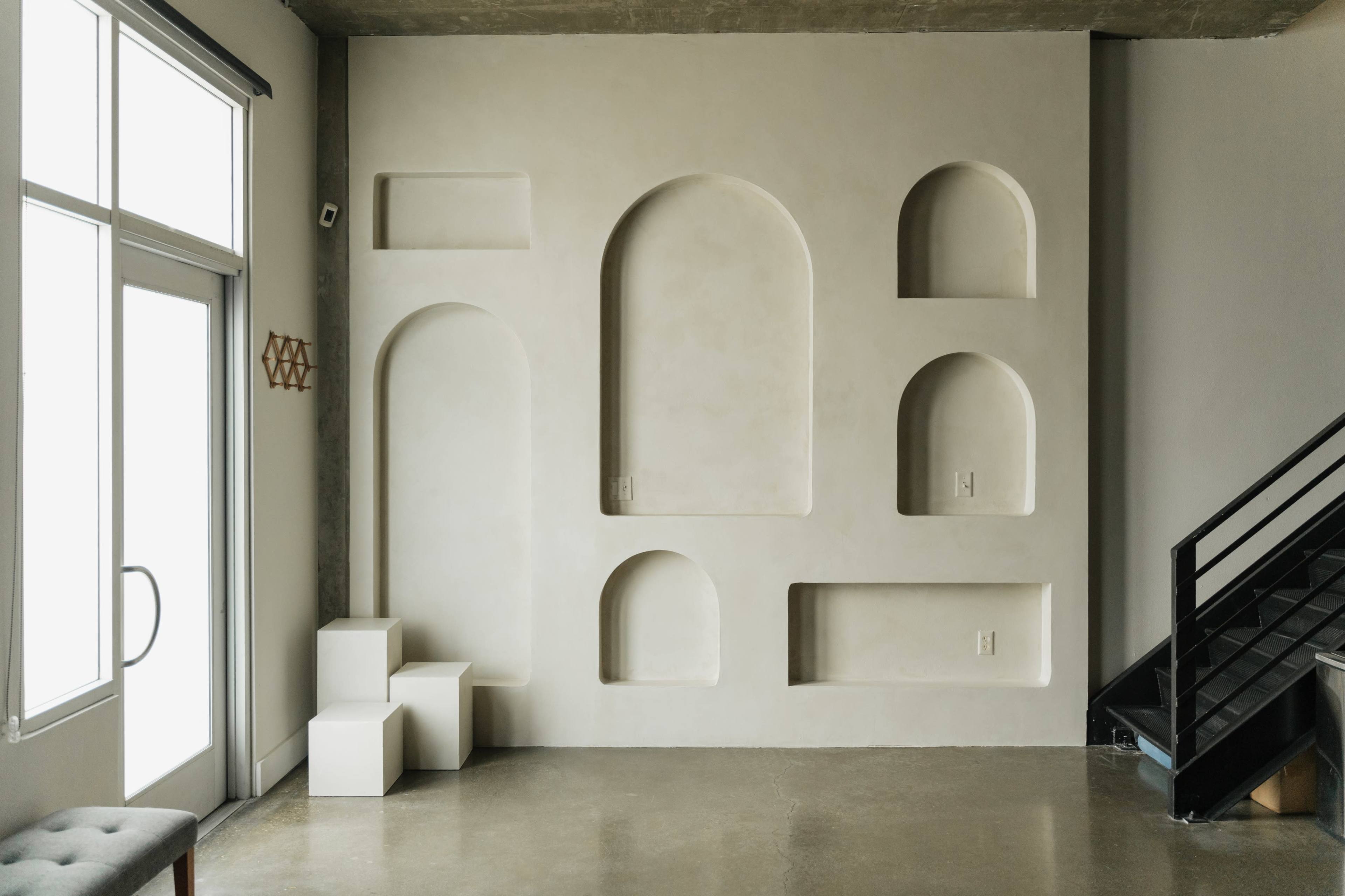 The image shows a minimalist interior with a textured wall featuring arched alcoves and a set of white cubic pedestals beside a glass door and staircase.