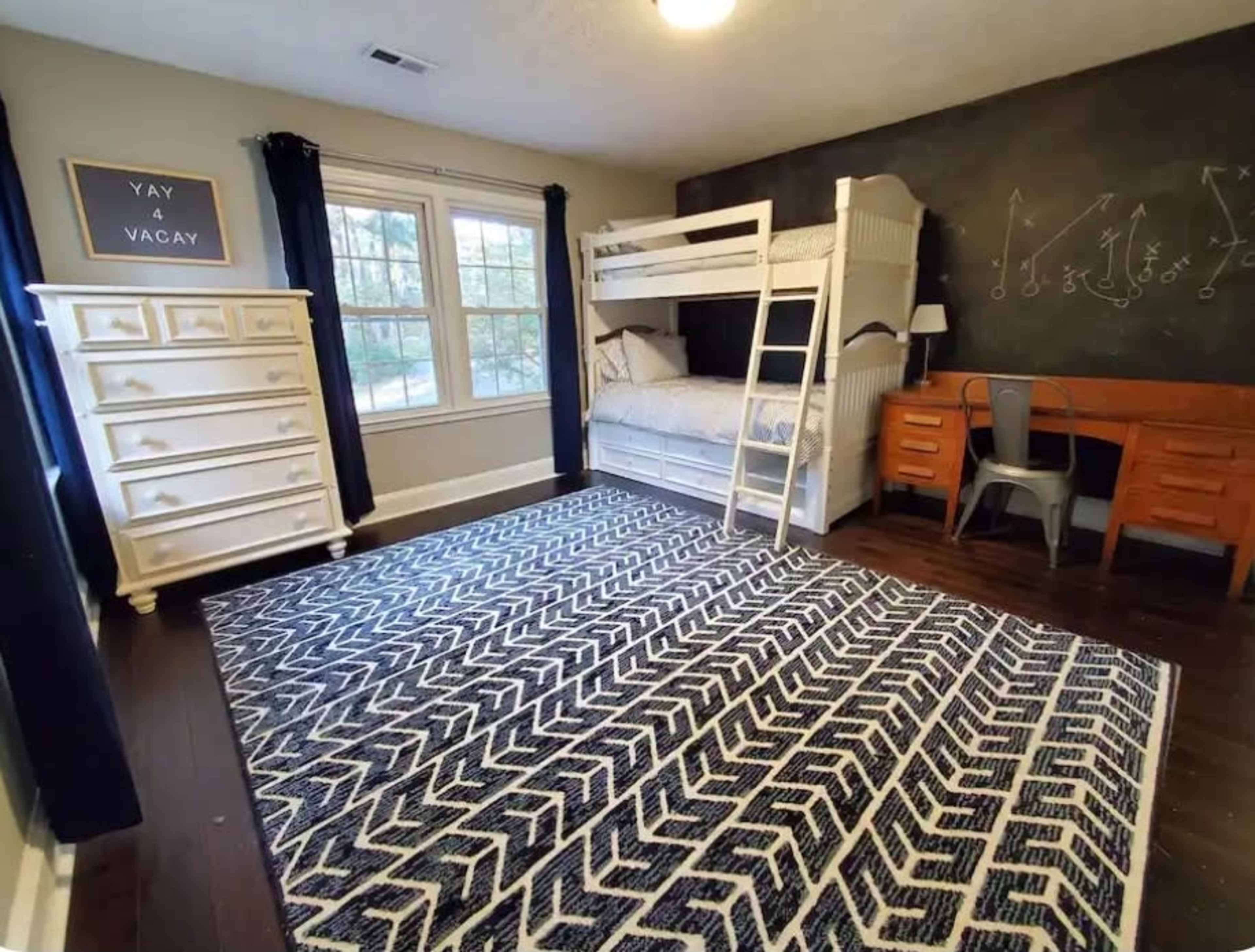 The image shows a room featuring a bunk bed, a wooden desk with two chairs, a white dresser, and a patterned rug on dark wooden flooring.