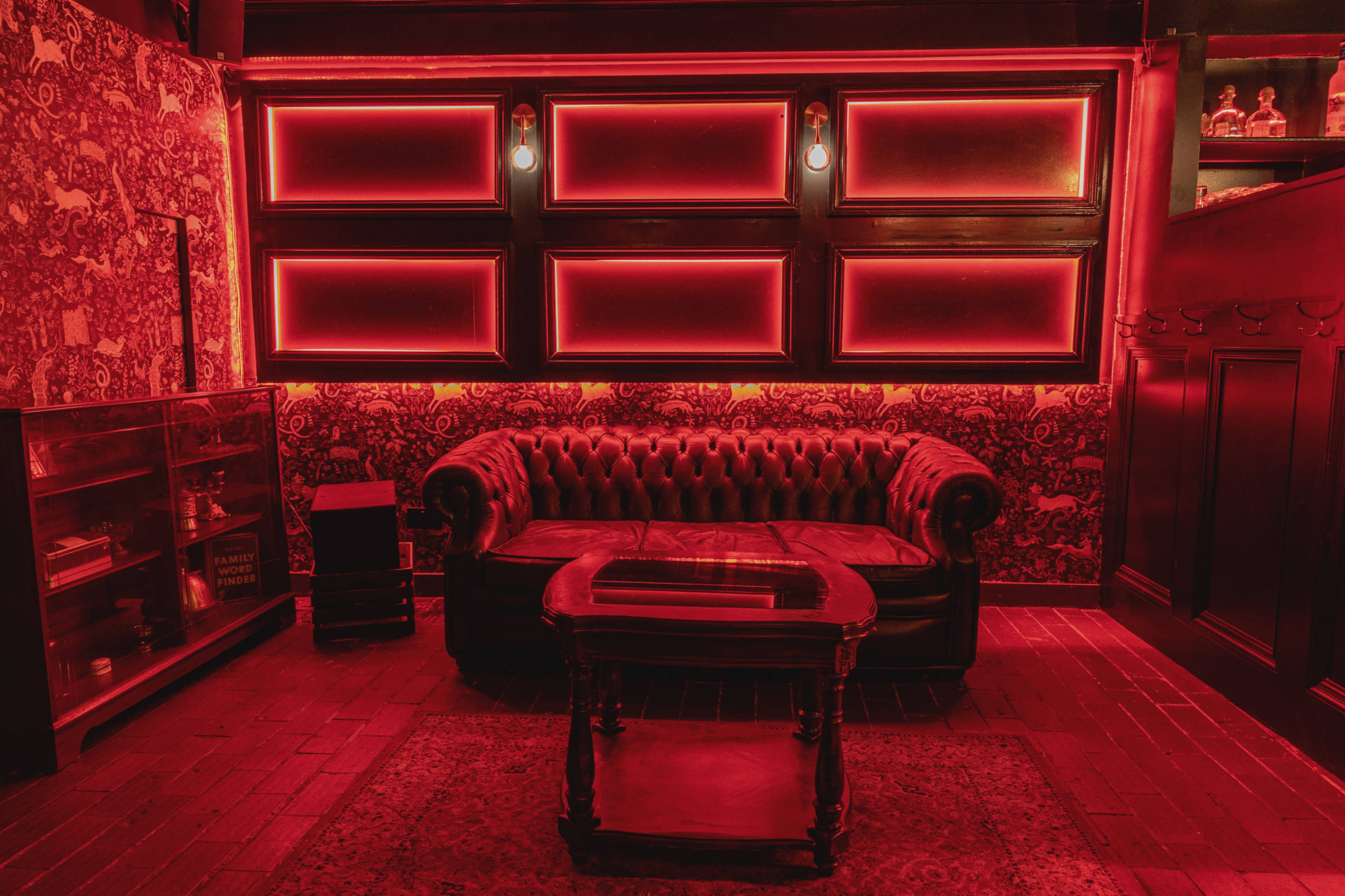 A vintage-style room features a red-lit leather couch and a wooden table set against patterned wallpaper.