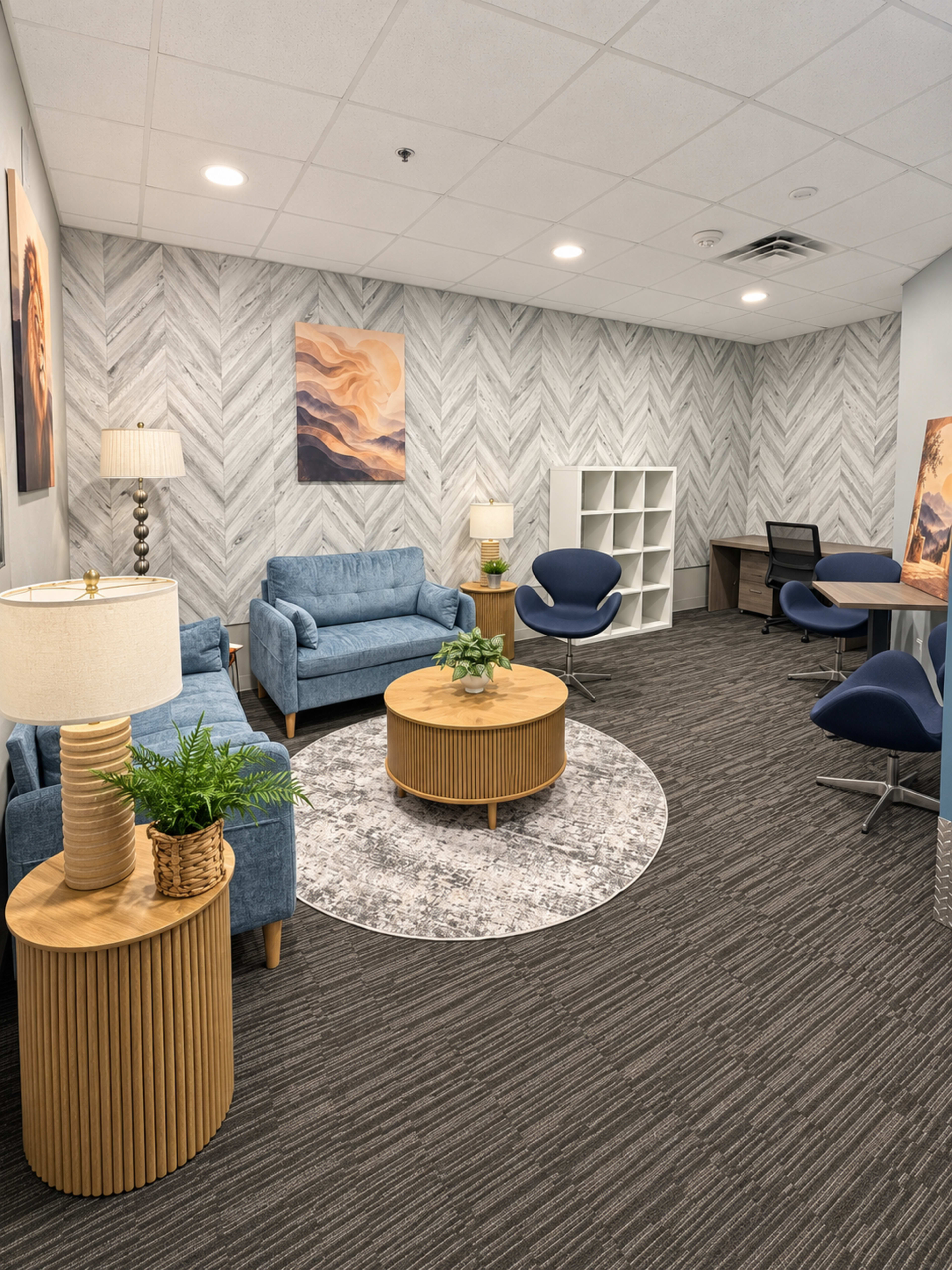 The image shows a modern waiting room with blue upholstered seating, a round wooden coffee table, and a small plant, all set against a chevron-patterned wall and complemented by a light gray carpet.