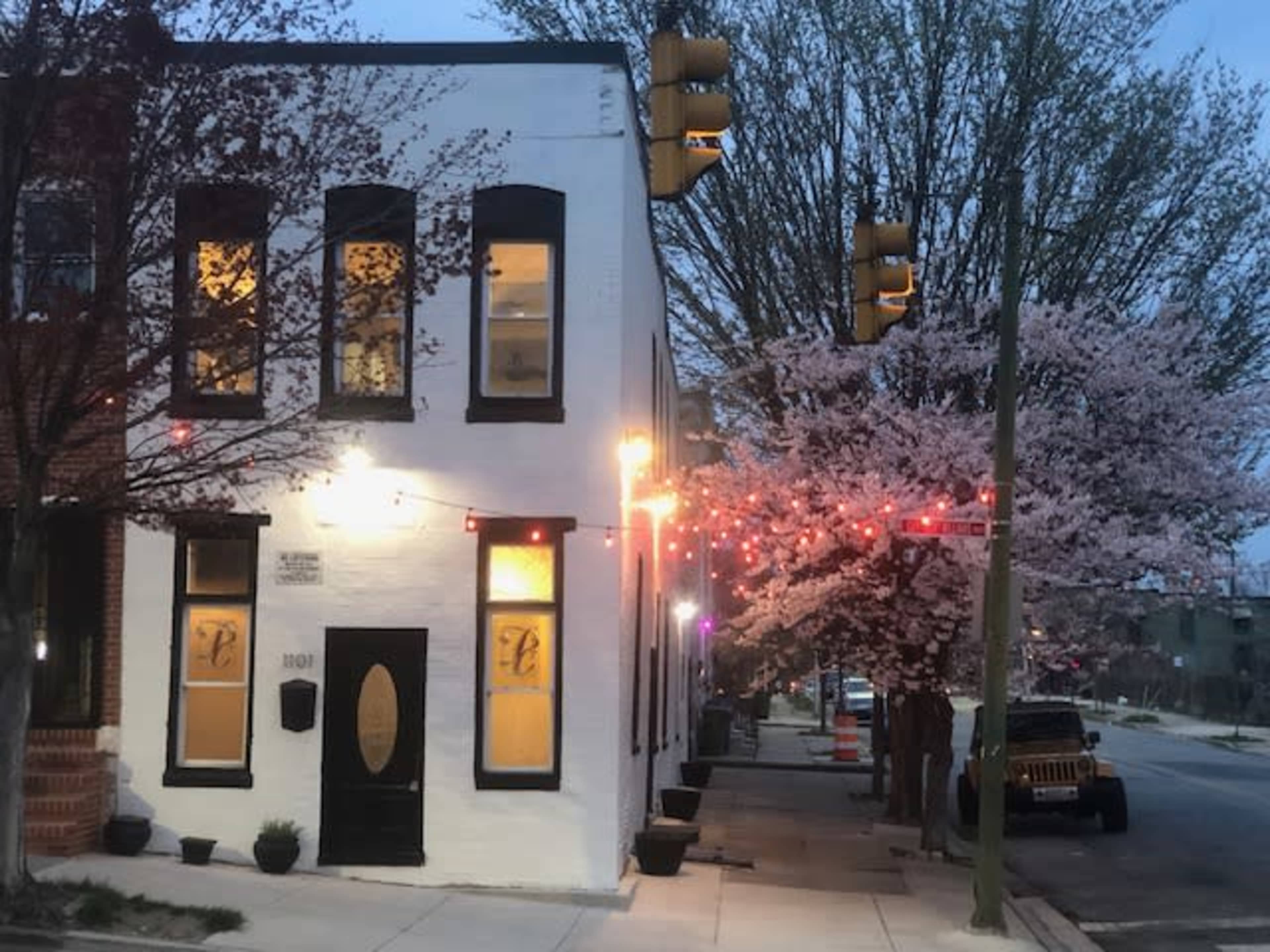 A white building with a round door stands at the corner of a street, illuminated by string lights and next to a tree in bloom.