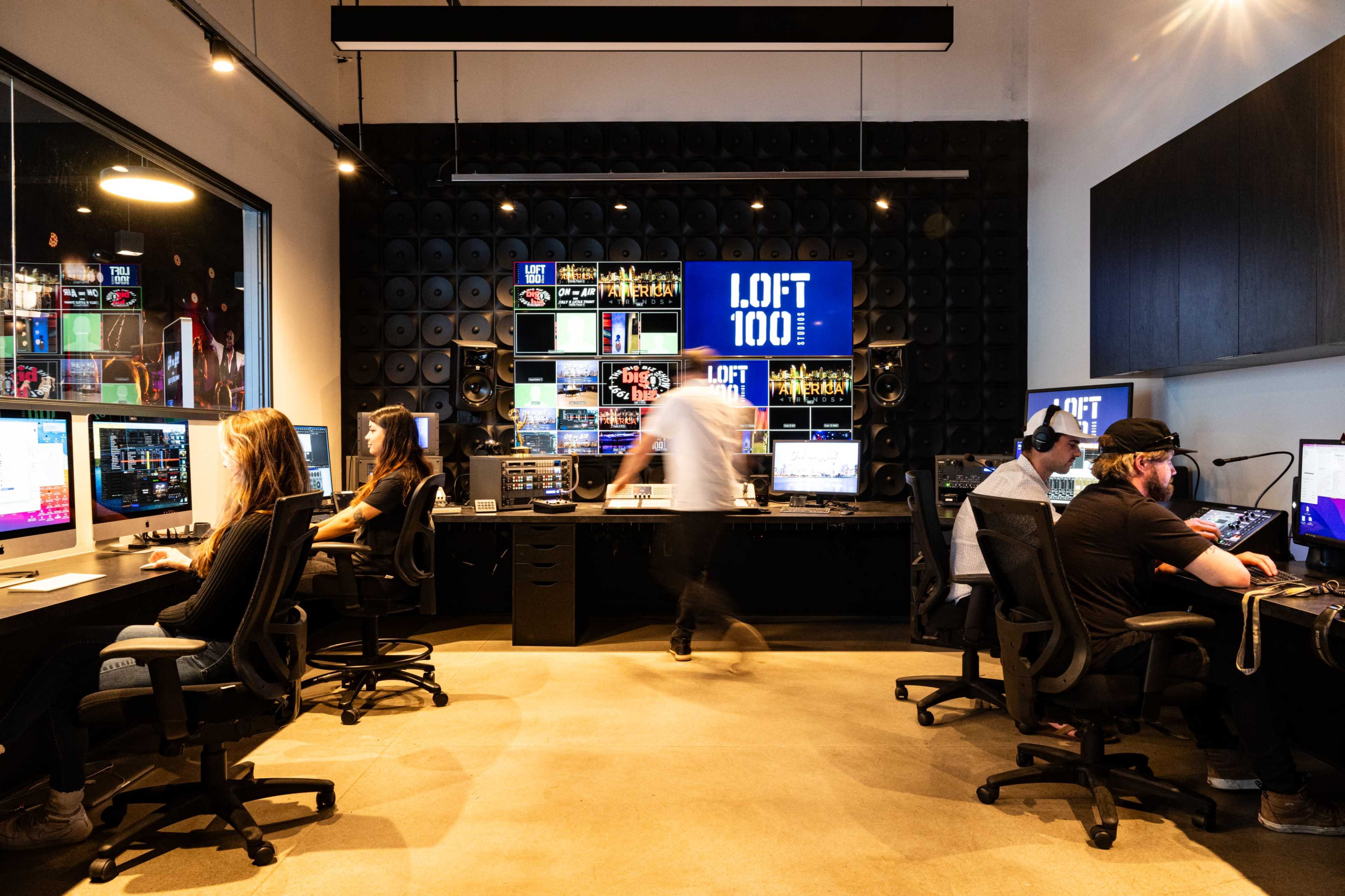 A modern media production studio features multiple workstations with computers and large screens displaying visuals, while individuals collaborate on projects.
