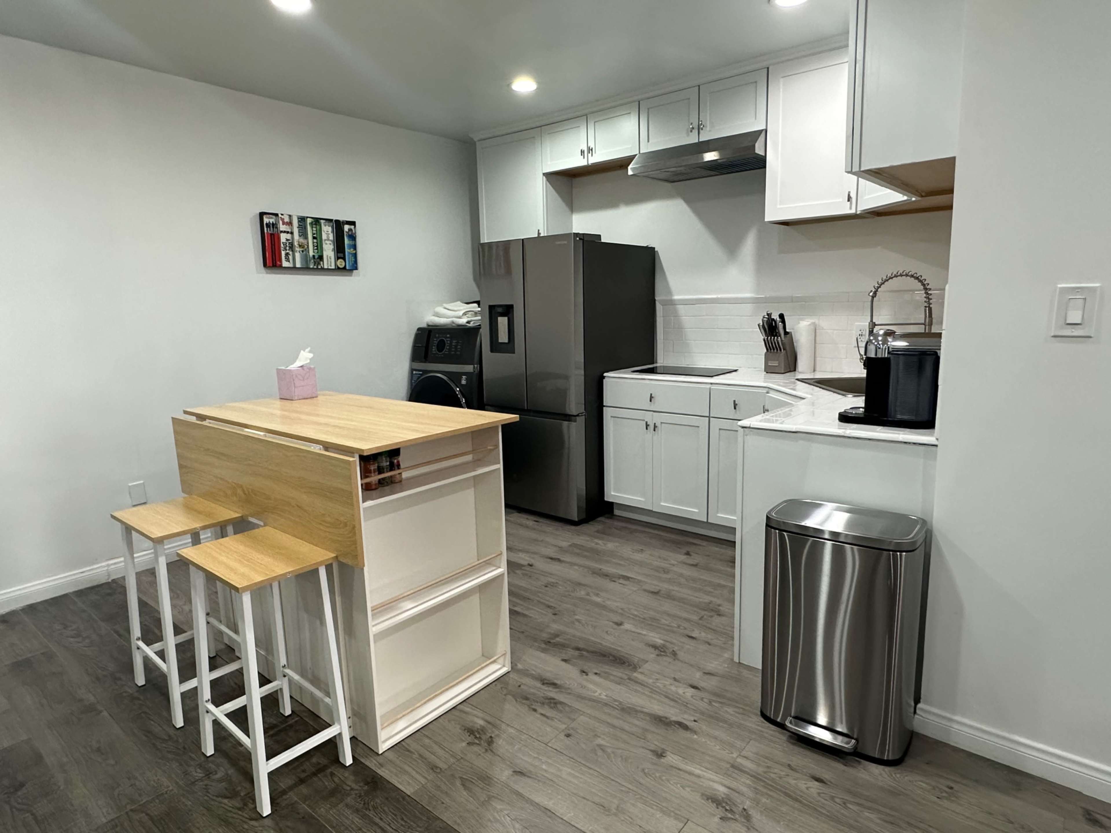 The image shows a modern kitchen with white cabinetry, stainless steel appliances, and a small wooden island with two stools.