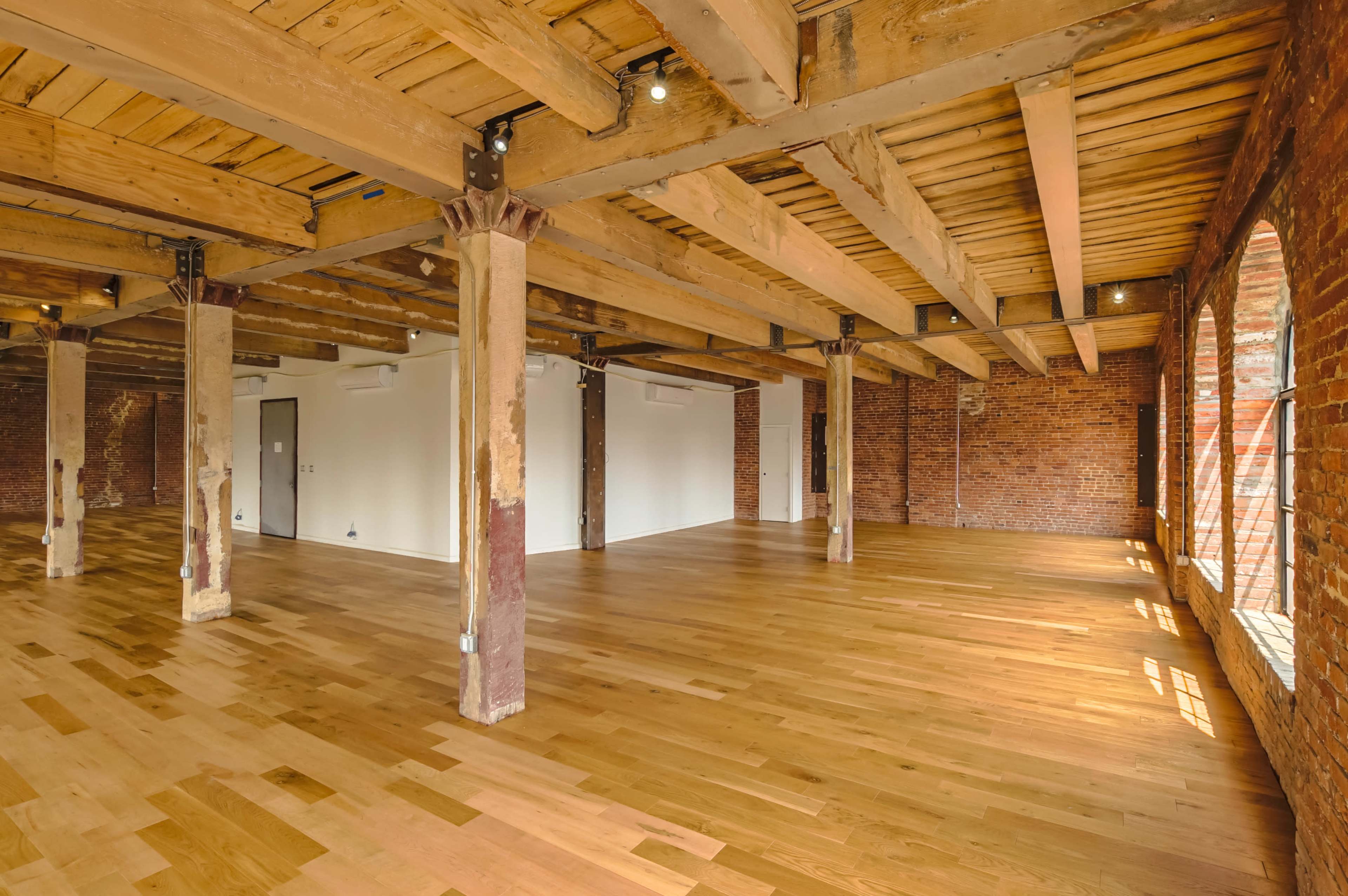 The image shows a spacious interior with wooden floors, exposed brick walls, and wooden beam ceilings, featuring several columns throughout the area.