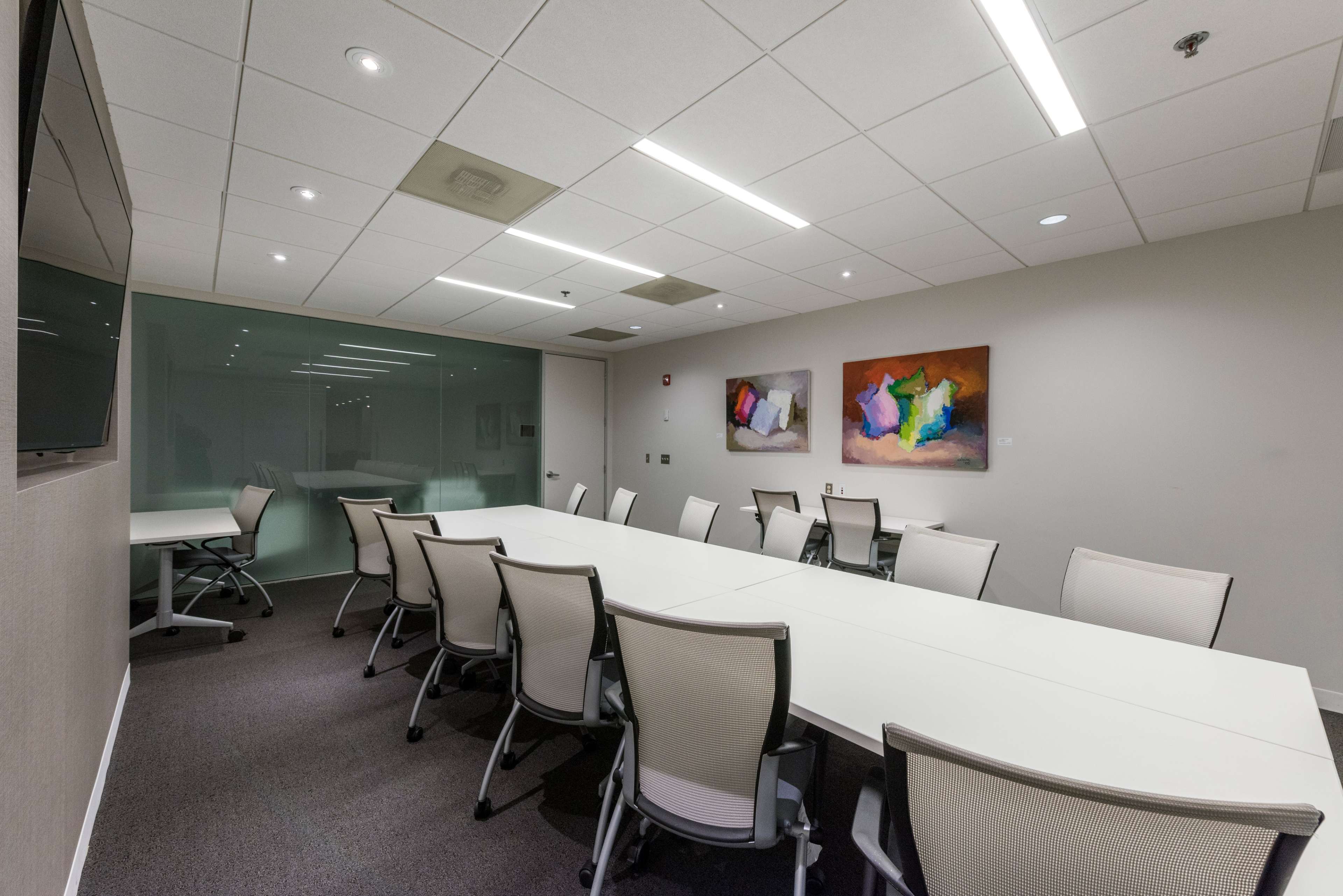 A conference room features a long white table surrounded by ergonomic chairs, with two colorful abstract paintings on the wall.