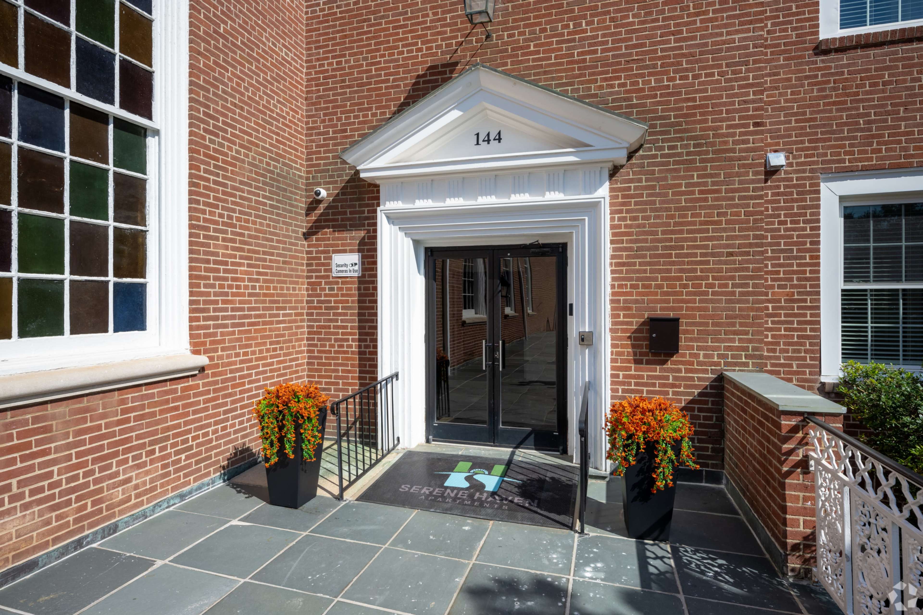 The entrance to a brick building features double glass doors, flanked by two potted orange flower arrangements, and a welcome mat displaying the word "Serenity."