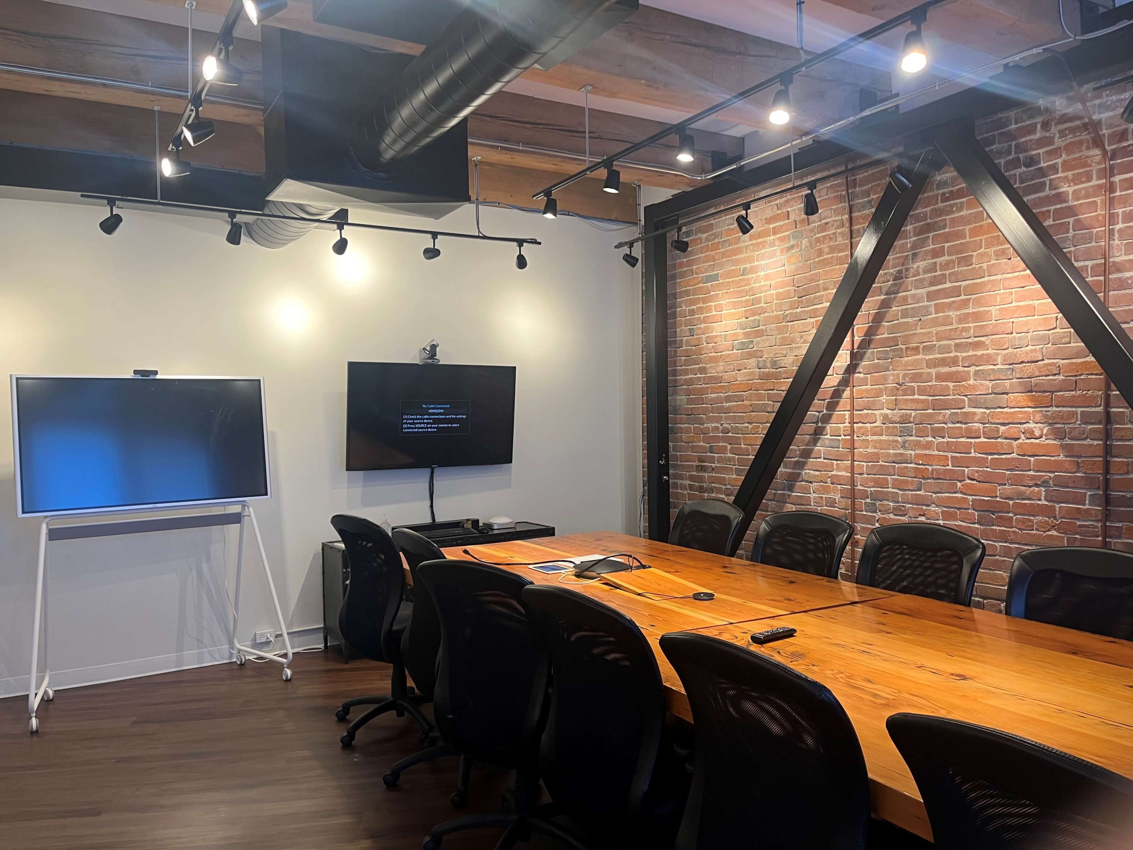 A modern conference room with a long wooden table, black ergonomic chairs, and two wall-mounted screens surrounded by exposed brick walls and overhead lighting.