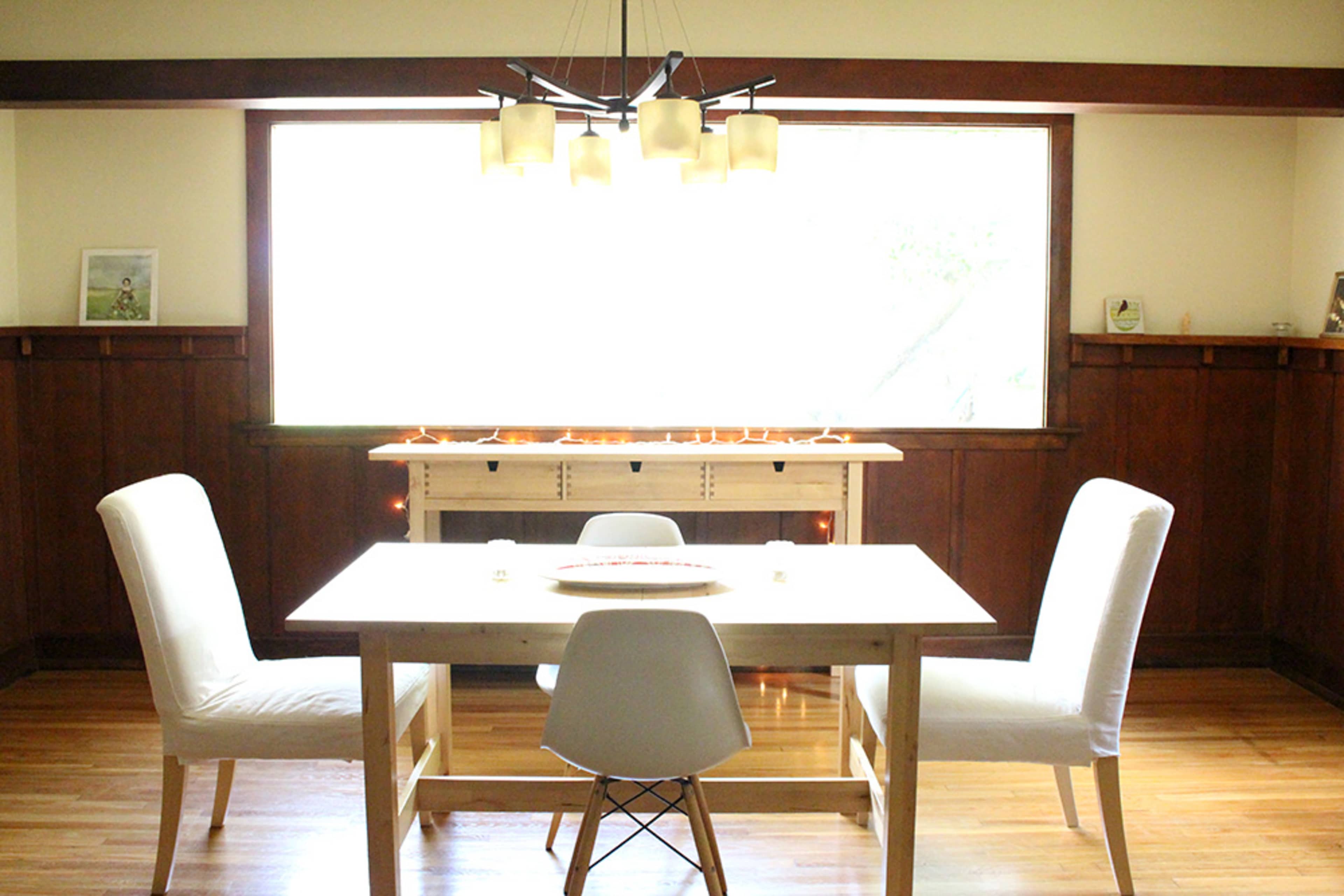 A simple dining area features a wooden table surrounded by four chairs, illuminated by a hanging light above and a large window letting in natural light.