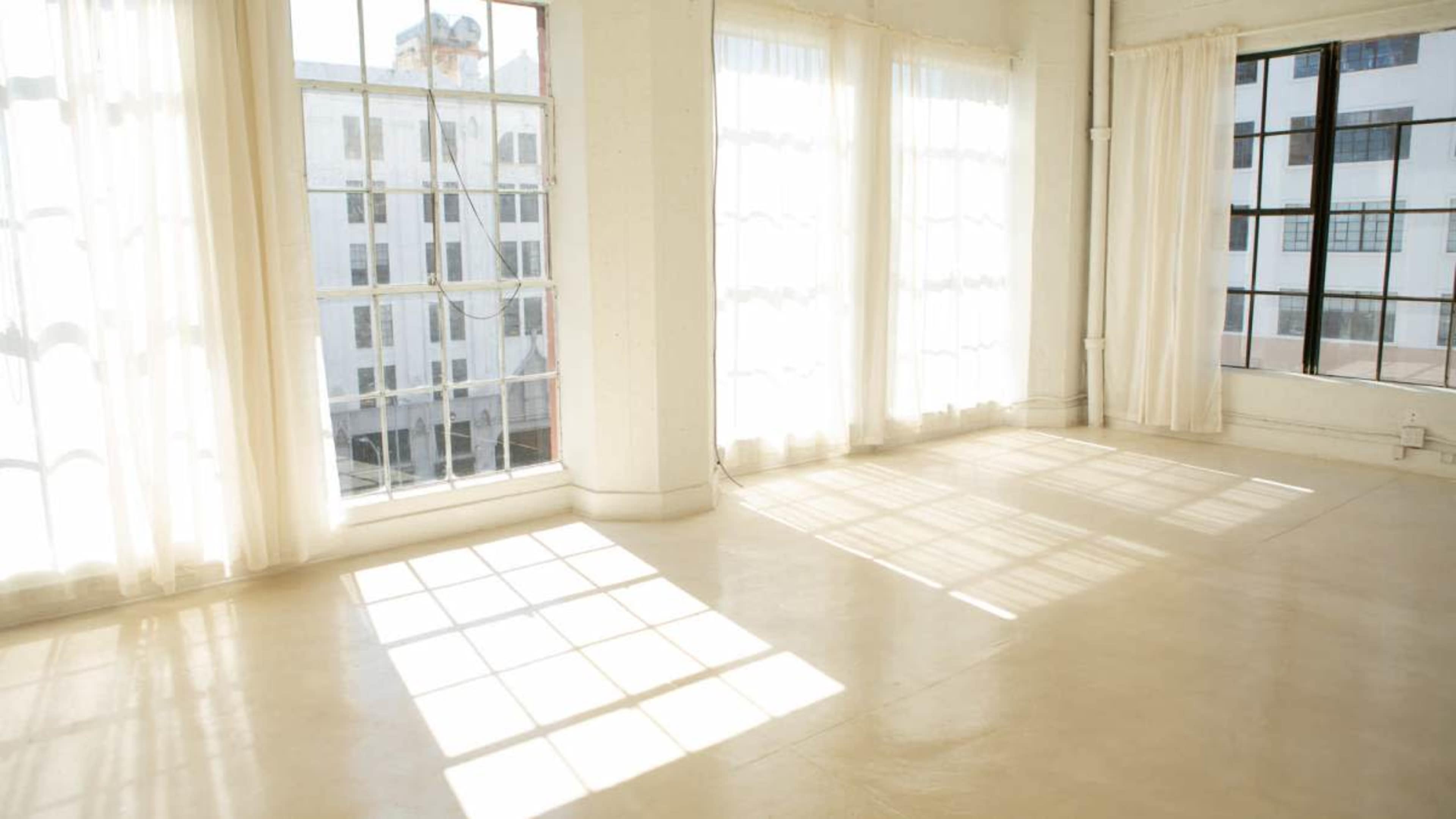 A bright, spacious room features large windows with white curtains, allowing sunlight to cast shadows on the light-colored floor.