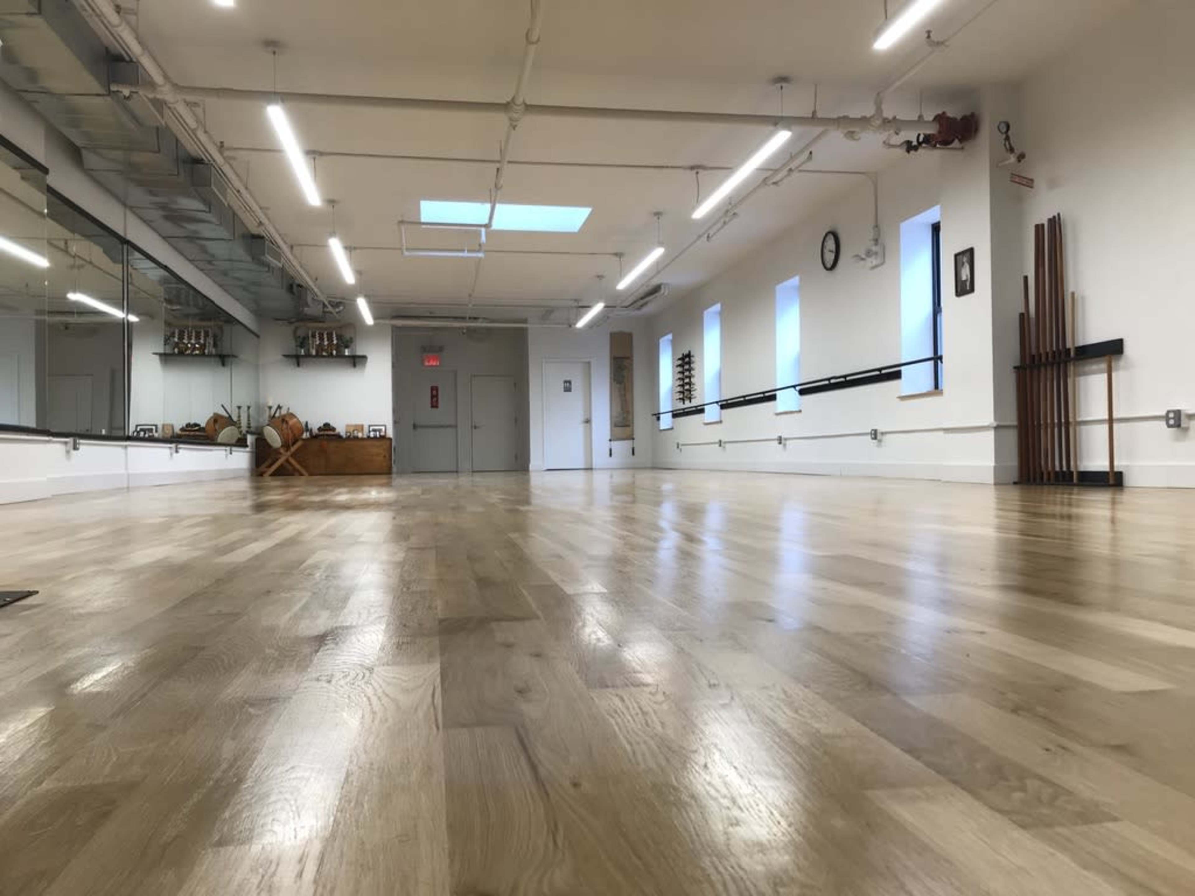 The image shows a spacious, well-lit dance studio with a wooden floor, mirrors, and a variety of exercise equipment against the wall.