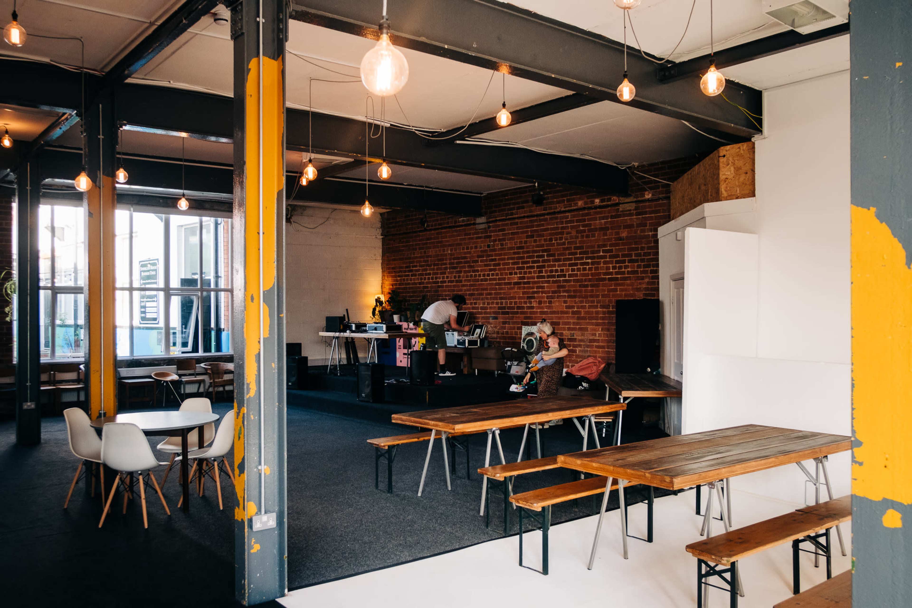 A spacious café with wooden tables, industrial-style lighting, and a small stage area at the back where a person is working.