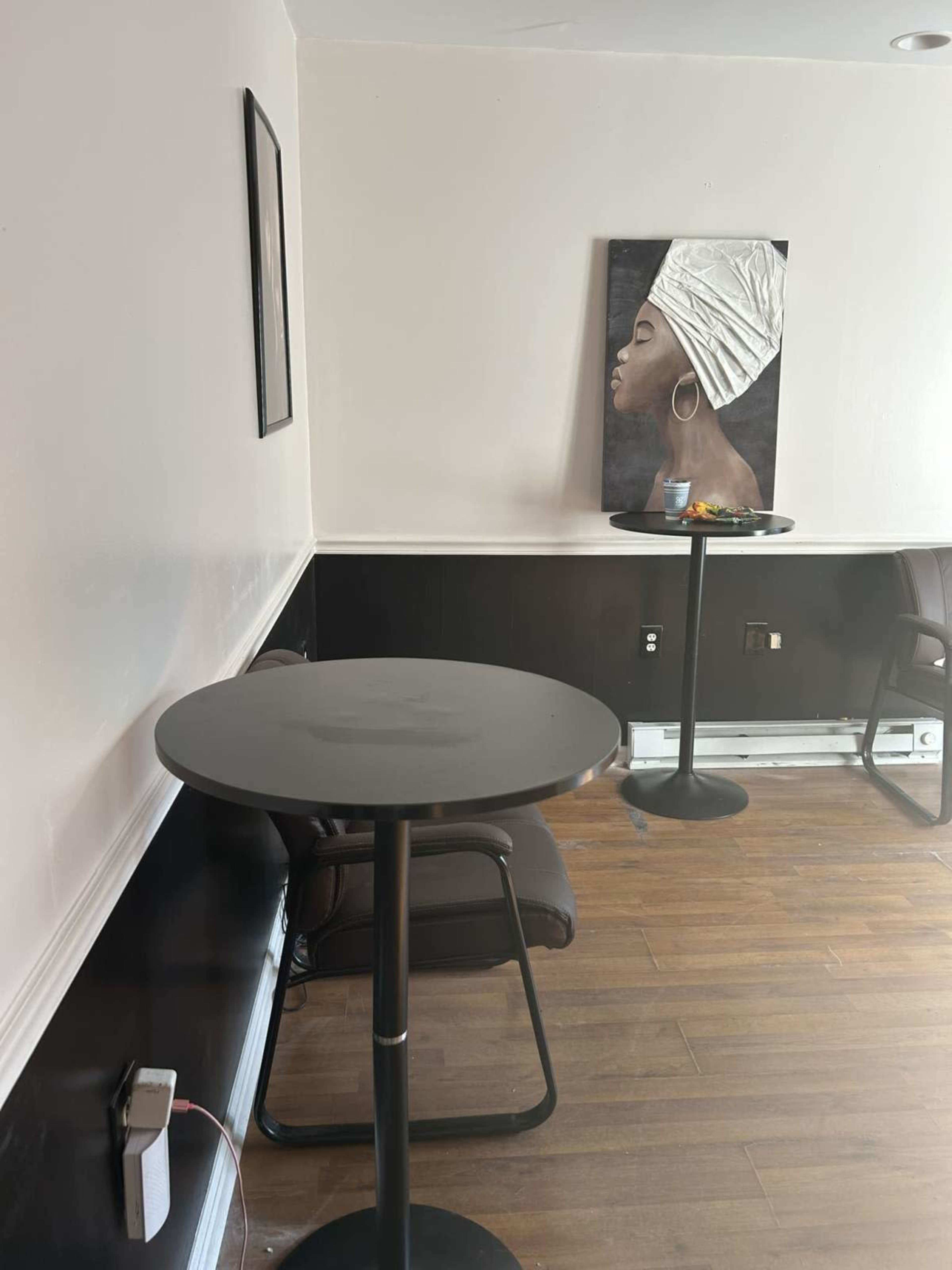 The image shows a minimalist café interior featuring two small black tables and a wall adorned with a portrait of a woman wearing a headwrap.