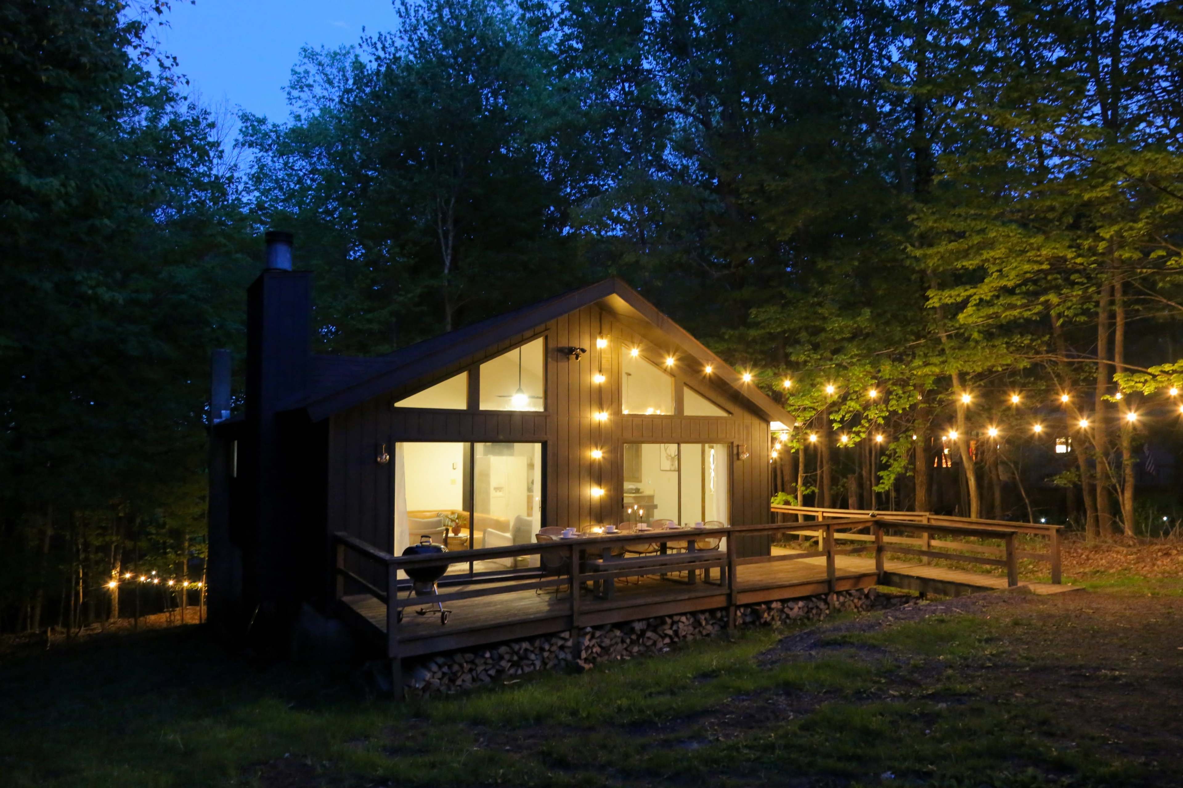 A cabin with large windows and string lights is situated in a wooded area at dusk.