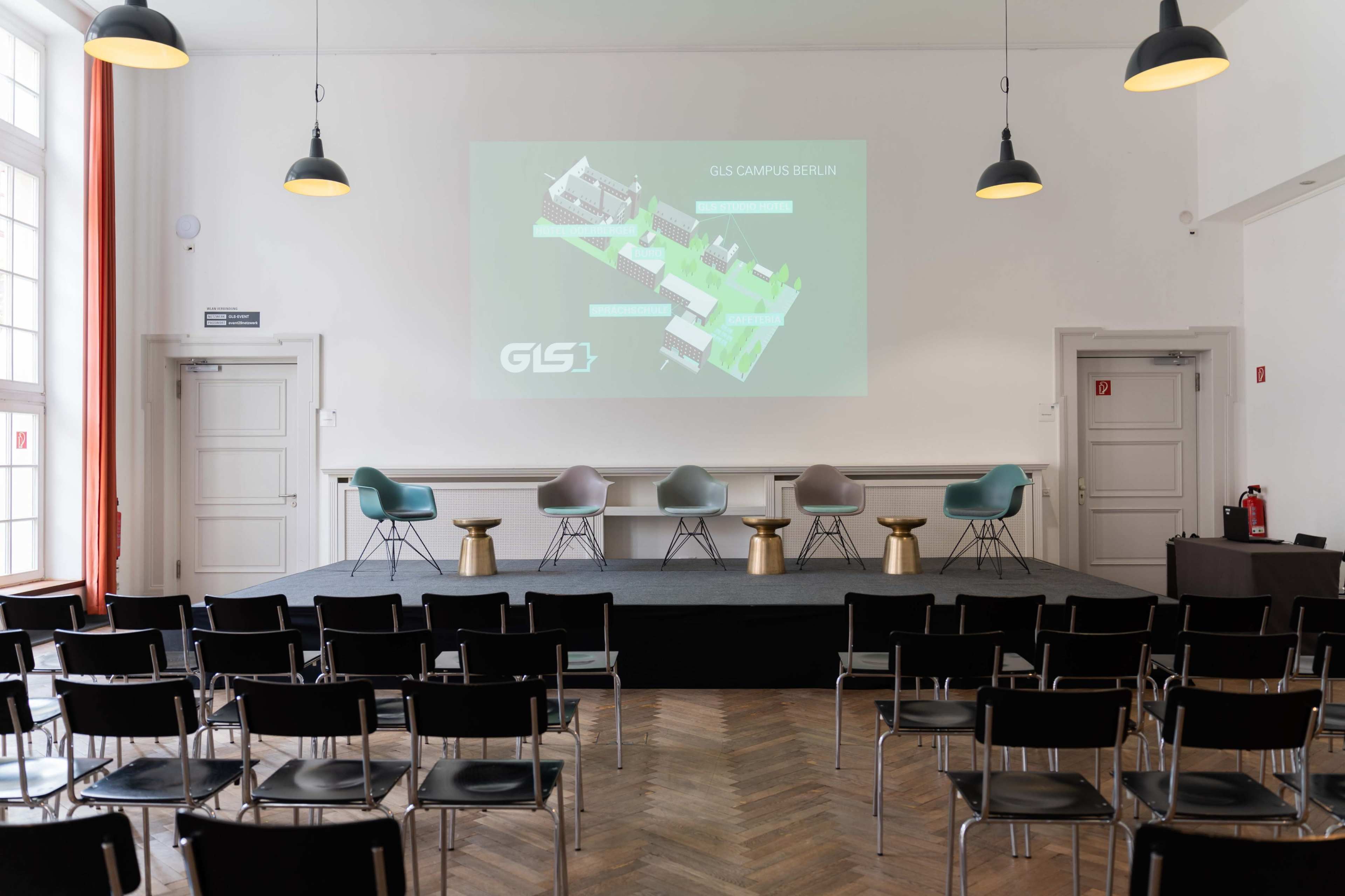 A conference room is set up with a presentation screen, panel seating, and rows of empty chairs facing the stage.