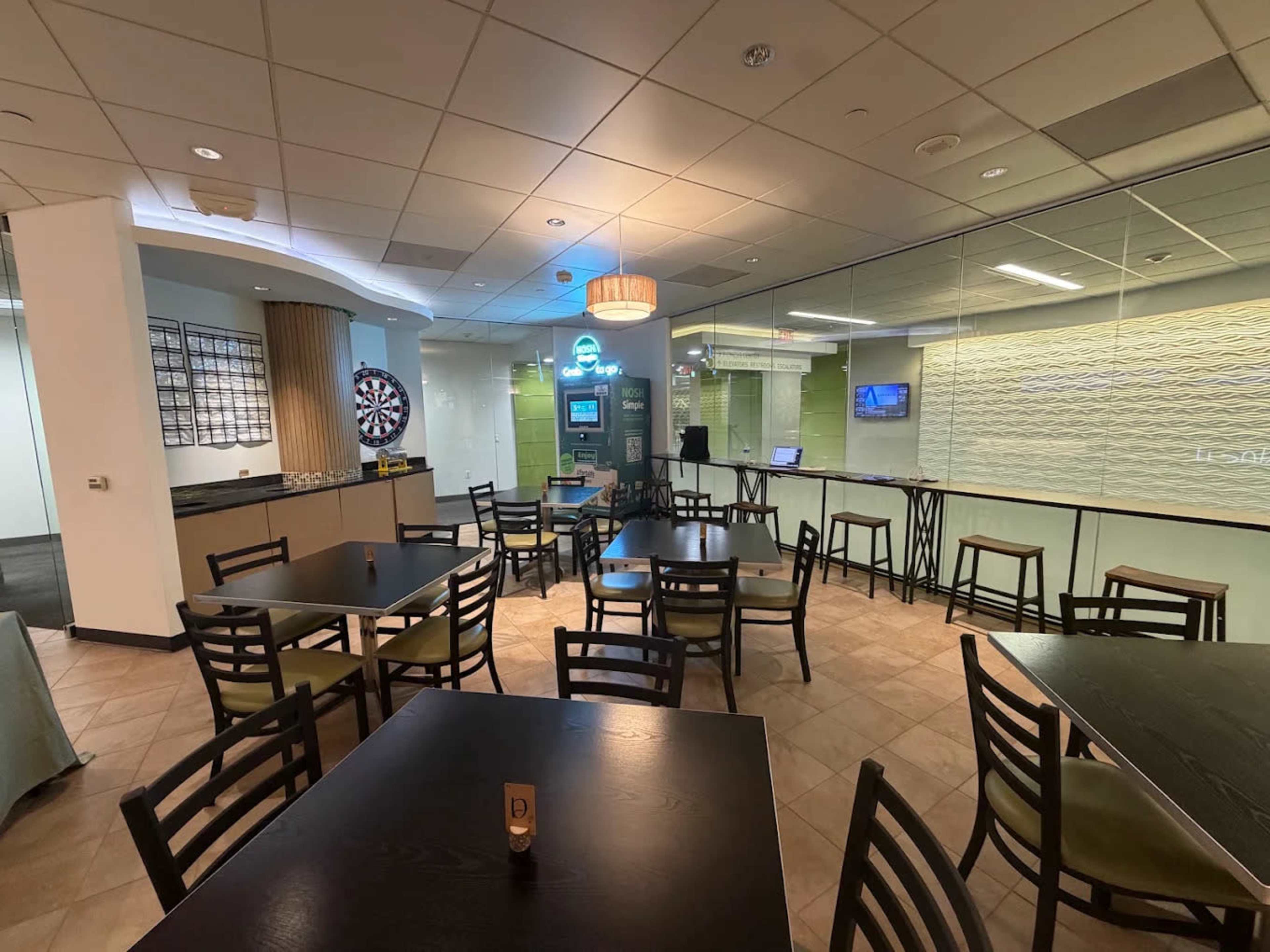 The image shows a modern cafe or lounge area with tables and chairs, a dartboard in the background, and a glass partition separating it from another space.