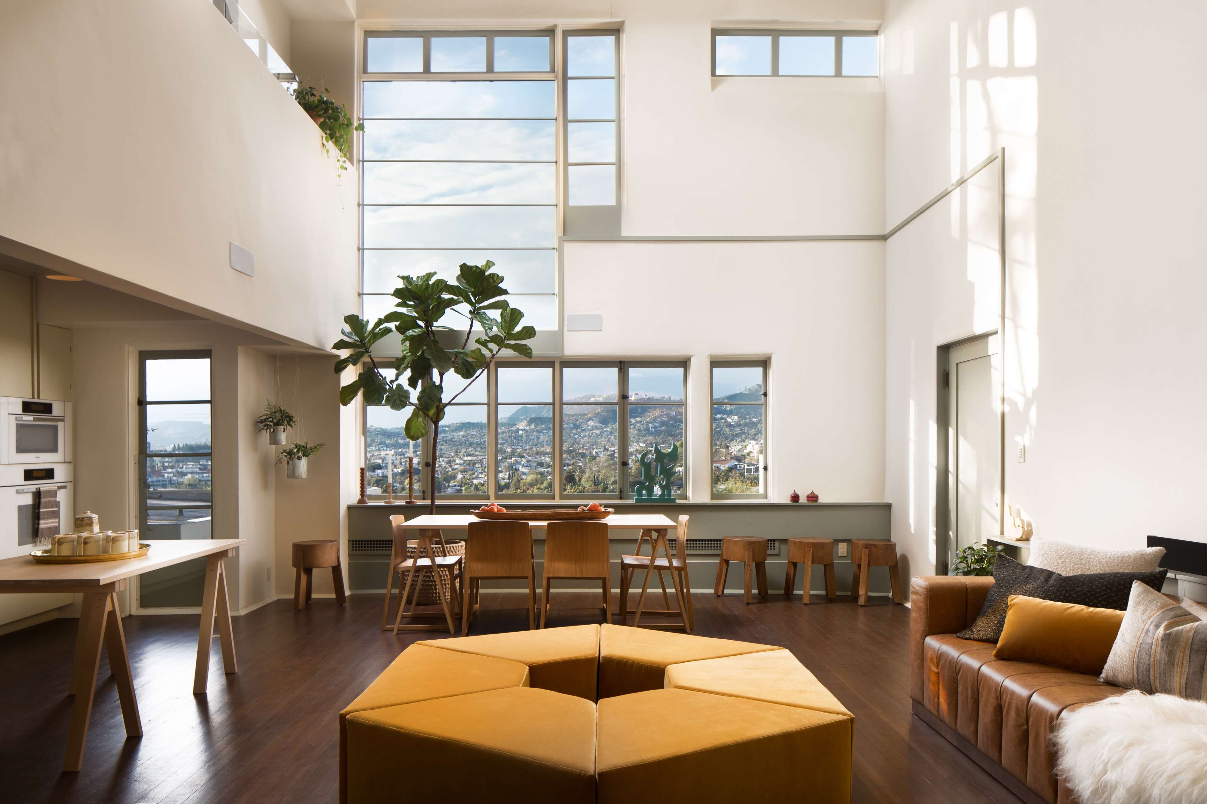 A spacious modern living area with large windows, a dining table, and a sectional couch, all featuring minimalist furnishings and a view of the outdoors.